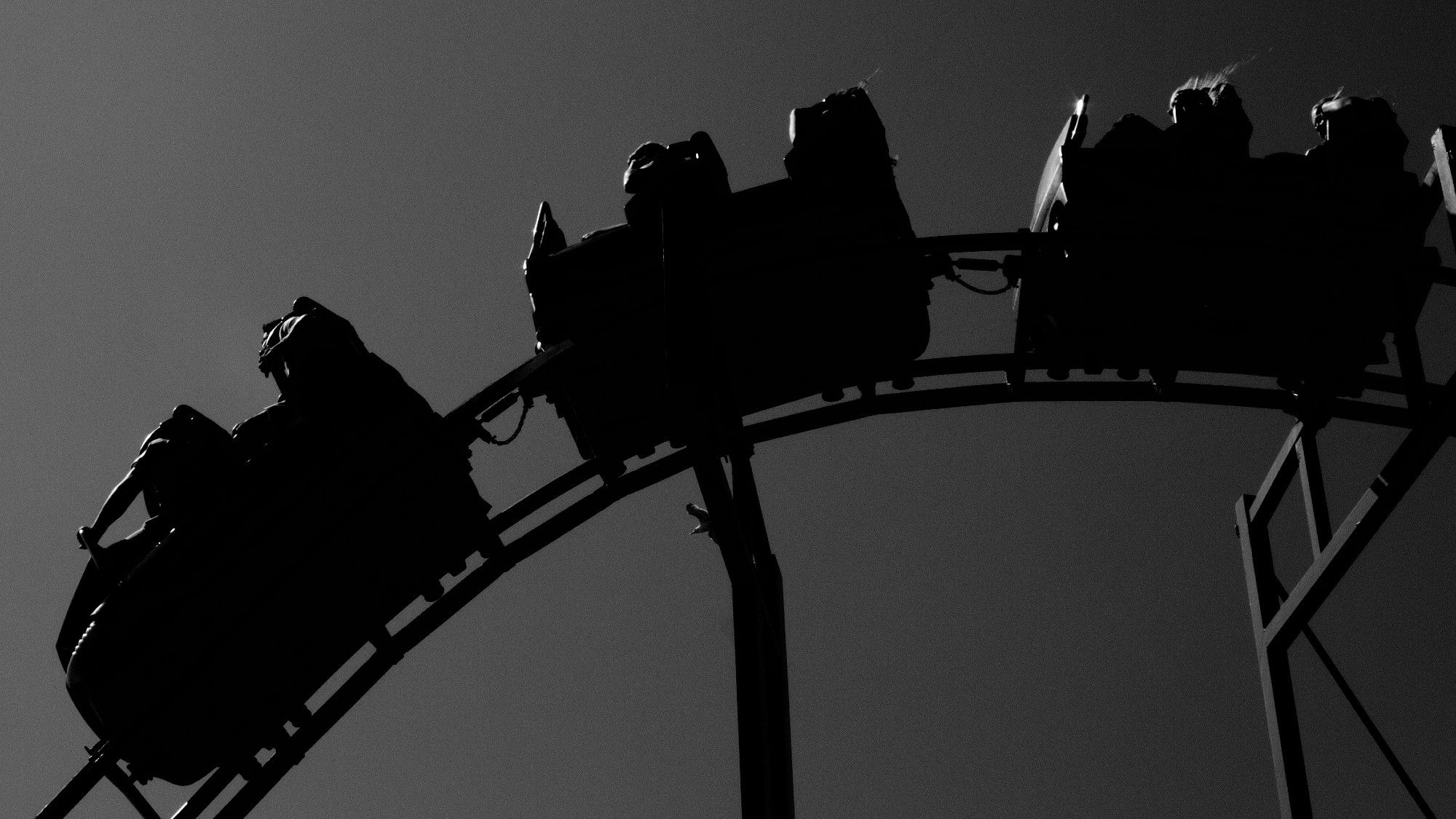 A black and white photo of a rollercoaster. The rollercoaster is mostly black except for a few slight glimpses where the sunlight catches it