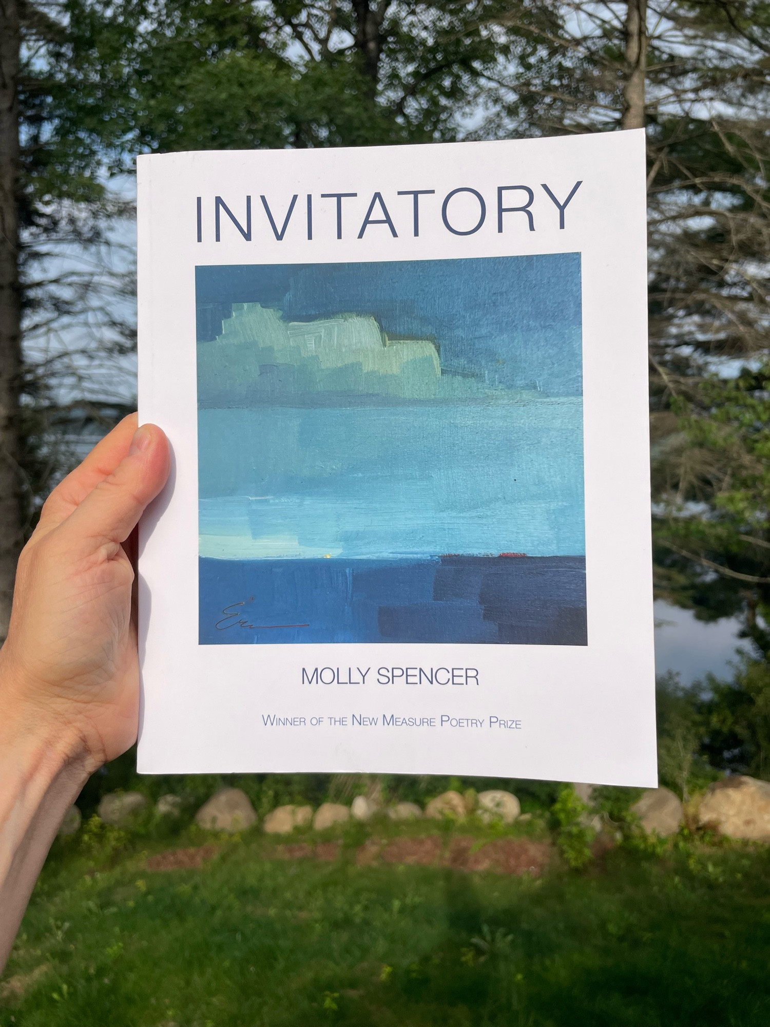 The cover of Invitatory by Molly Spencer has an image with wide fields of thick blue that are sky and cloud and water and sky and a sliver of land, all stacked up on one another. In this picture, my hand holds the book in front of one of my favorite bodies of water—Long Lake in the Adirondacks. 