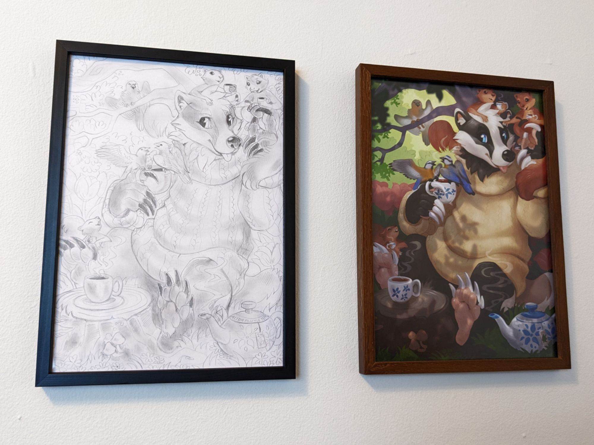 Photo of two pictures hanging side by side. Left is a pencil drawing in a black frame, of a badger in a sweater drinking tea with various forest animals. Right is the same drawing colored and shaded, in a dark brown frame.