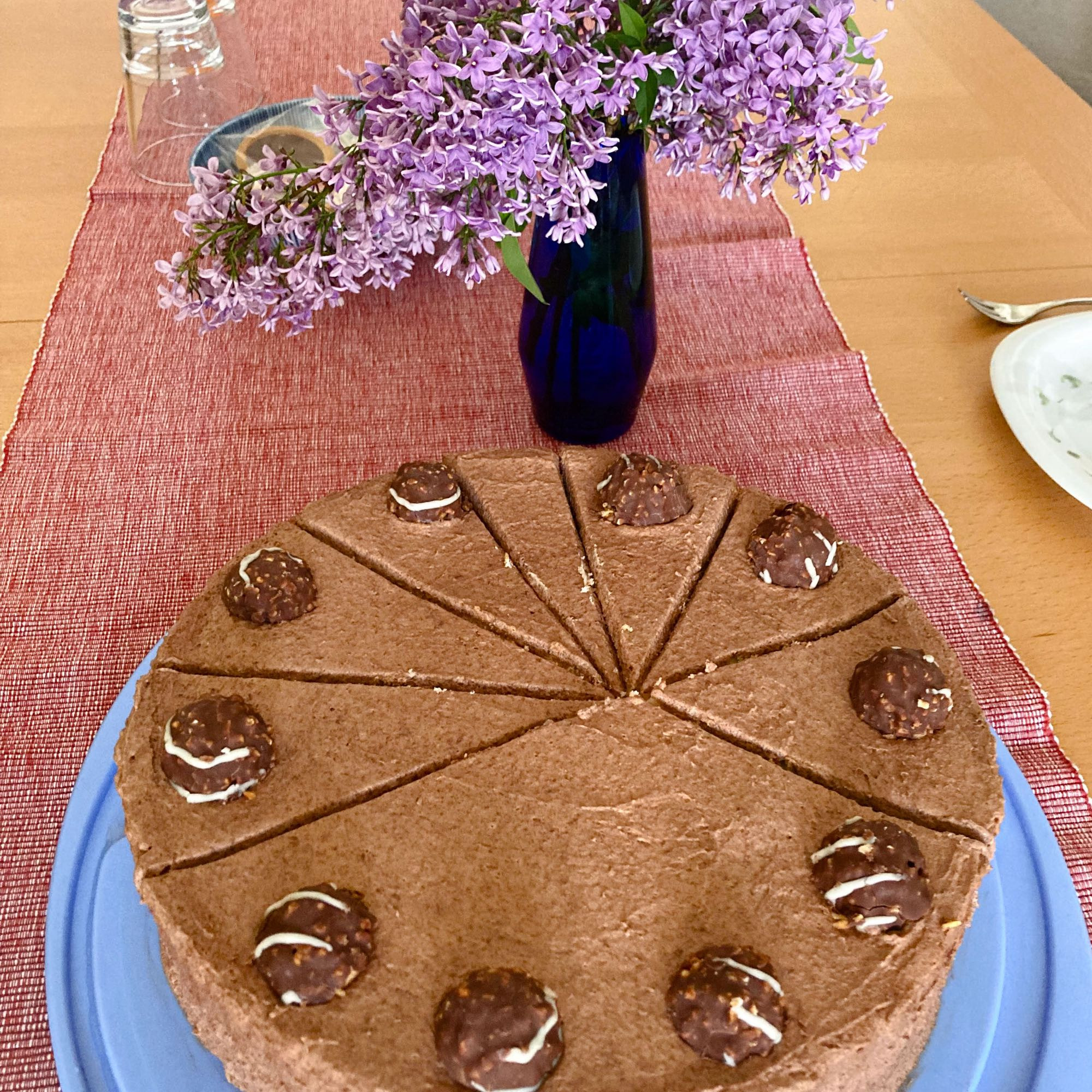 Eine mit Pralinen belegte Schokotorte, die in sehr unregelmäßig große Stücke geschnitten ist. Ein Teil ist noch ungeschnitten und hat so statt 1 Praline wie die anderen 4 Pralinen.
Auf dem Tisch steht noch eine Vase mit Fliederblüten.