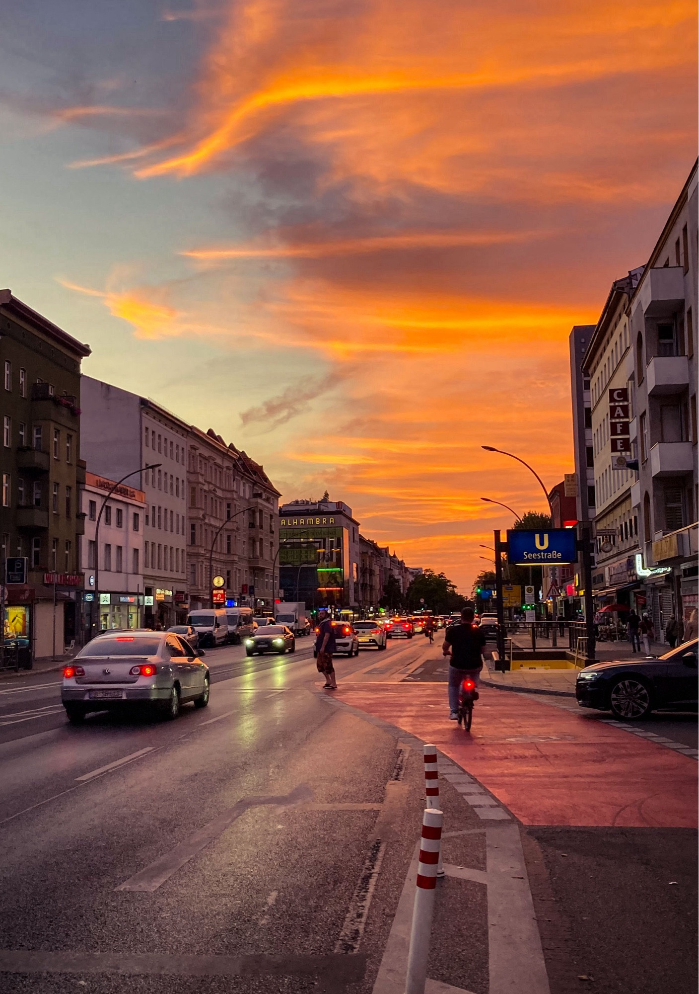 Straßenflucht mit dramatisch orangefarbenen Himmel. Auf der Straße fahren Autos, auf dem breiten Fahrradweg ein Fahrrad. An den Läden und dem Kino leuchten die Schilder bzw. Lichtbänder.