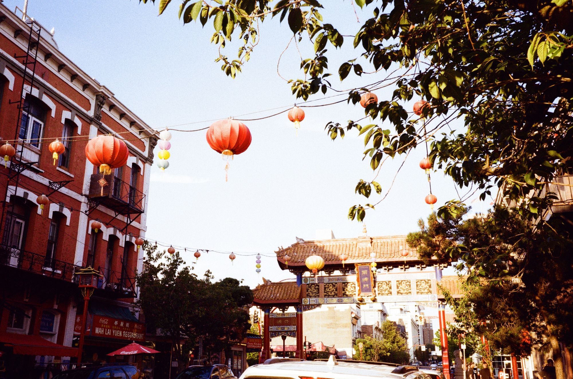 A film photograph of a city street with red and gold paifang in the background. In the foreground there are trees and red paper lanterns hanging from strings