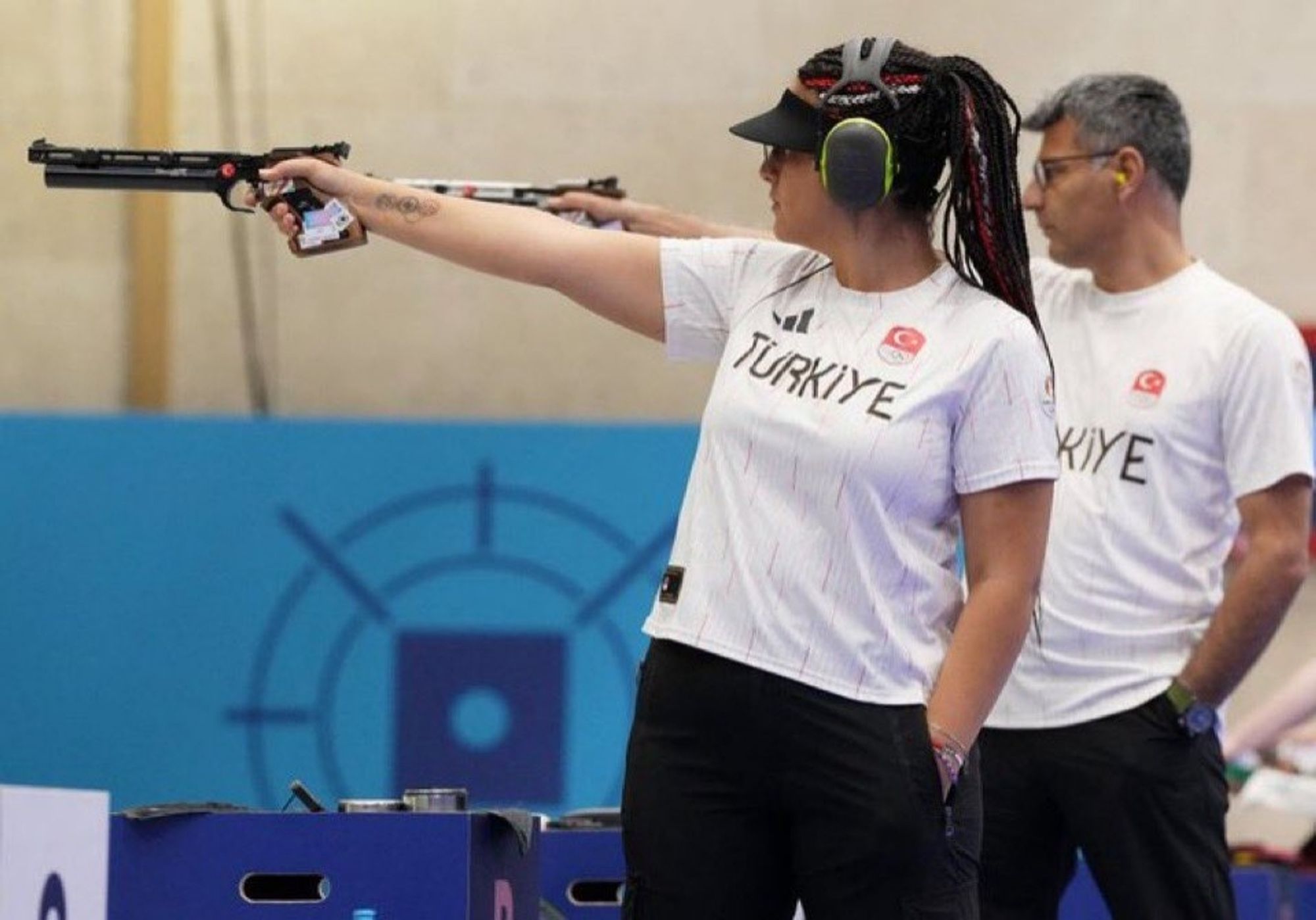 A woman and a man right behind her holding up their air pistols at their target during Olympic games. Both of them have their idle left hands in their pockets. They're wearing identical team uniforms of white tees and black slacks.