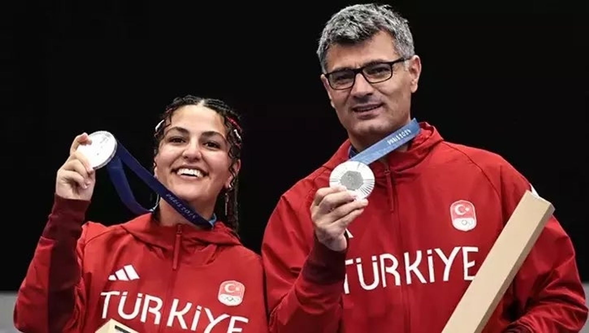 A brunette woman and a silver haired bespectacled man in red team track suits with Türkiye and Turkish flag in the front holding their silver medals.