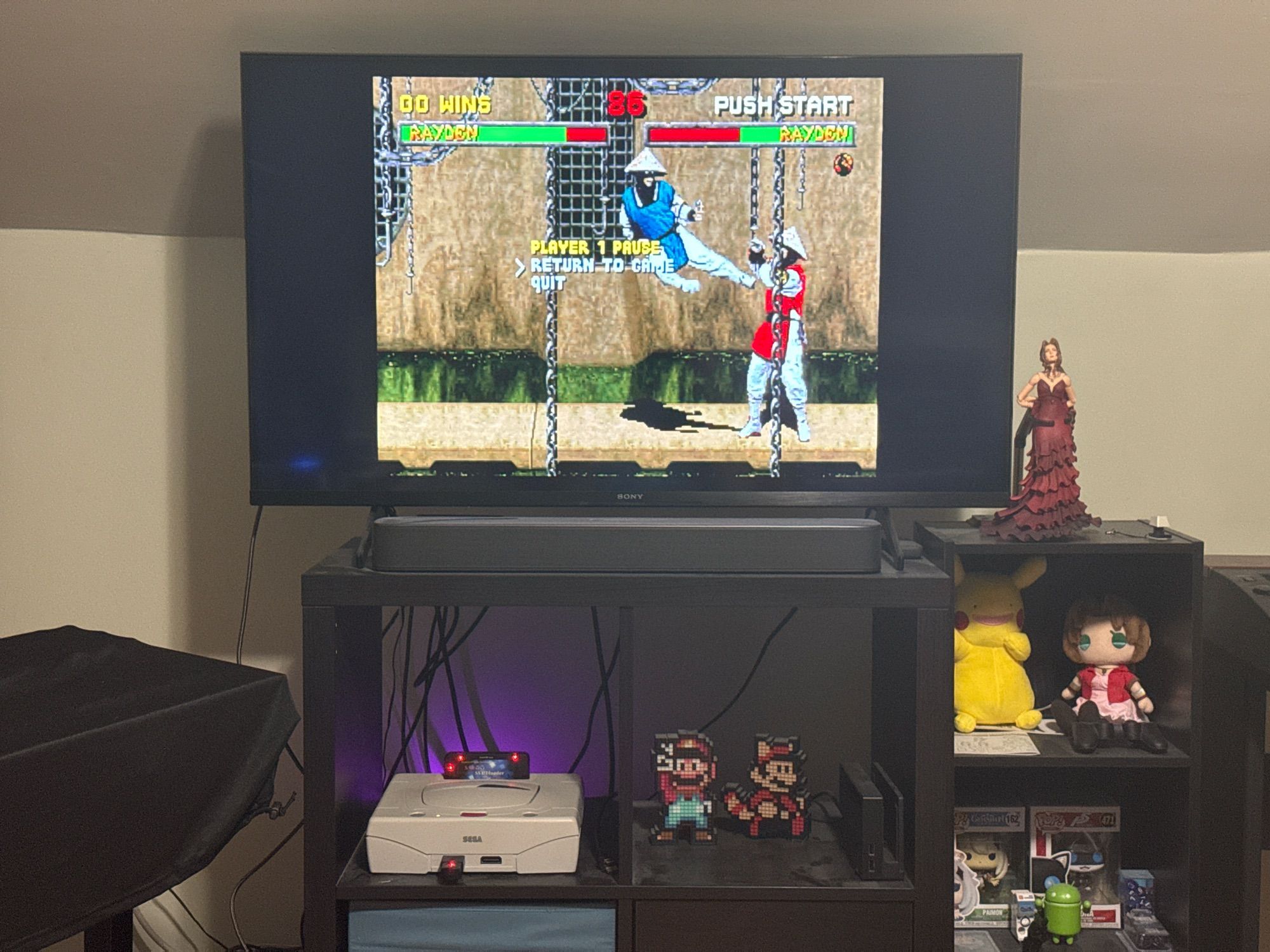 A TV showing Raiden jump kicking another Raiden in the Saturn version of Mortal Kombat 2. There are miscellaneous geeky toys in the room.