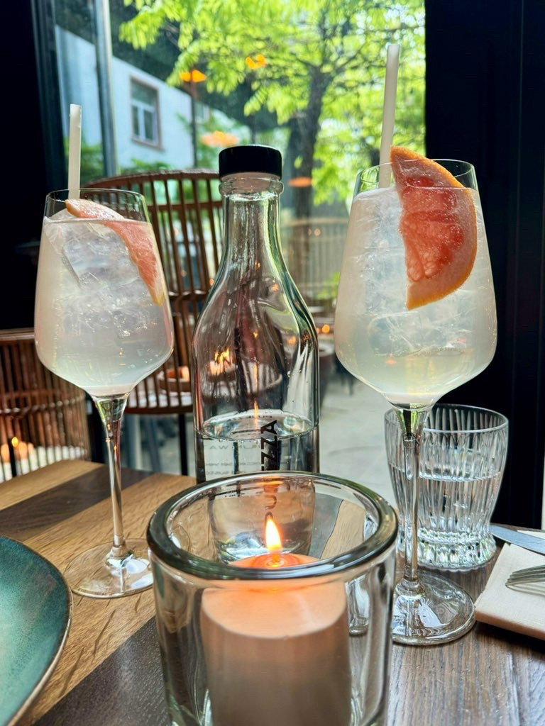 Zwei Gläser Grapefruit Spritz, eine Glasflasche Wasser, davor einen Kerze kn einem Glas. Im Hintergrund der begrünte Hof eines Restaurants