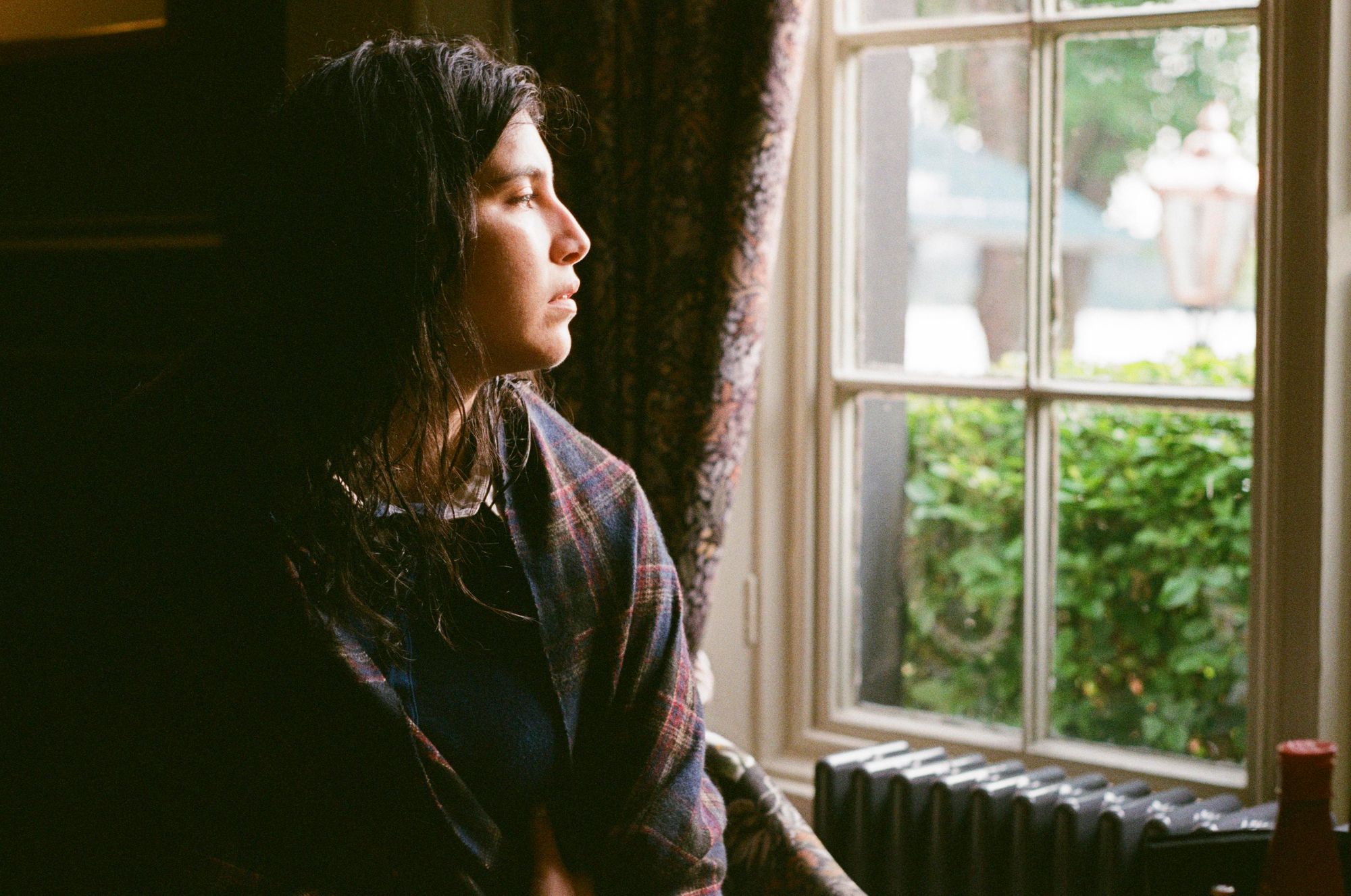 A woman with damp long black hair huddling a shawl around herself in front of a window with a view on a lake and a green hedge.