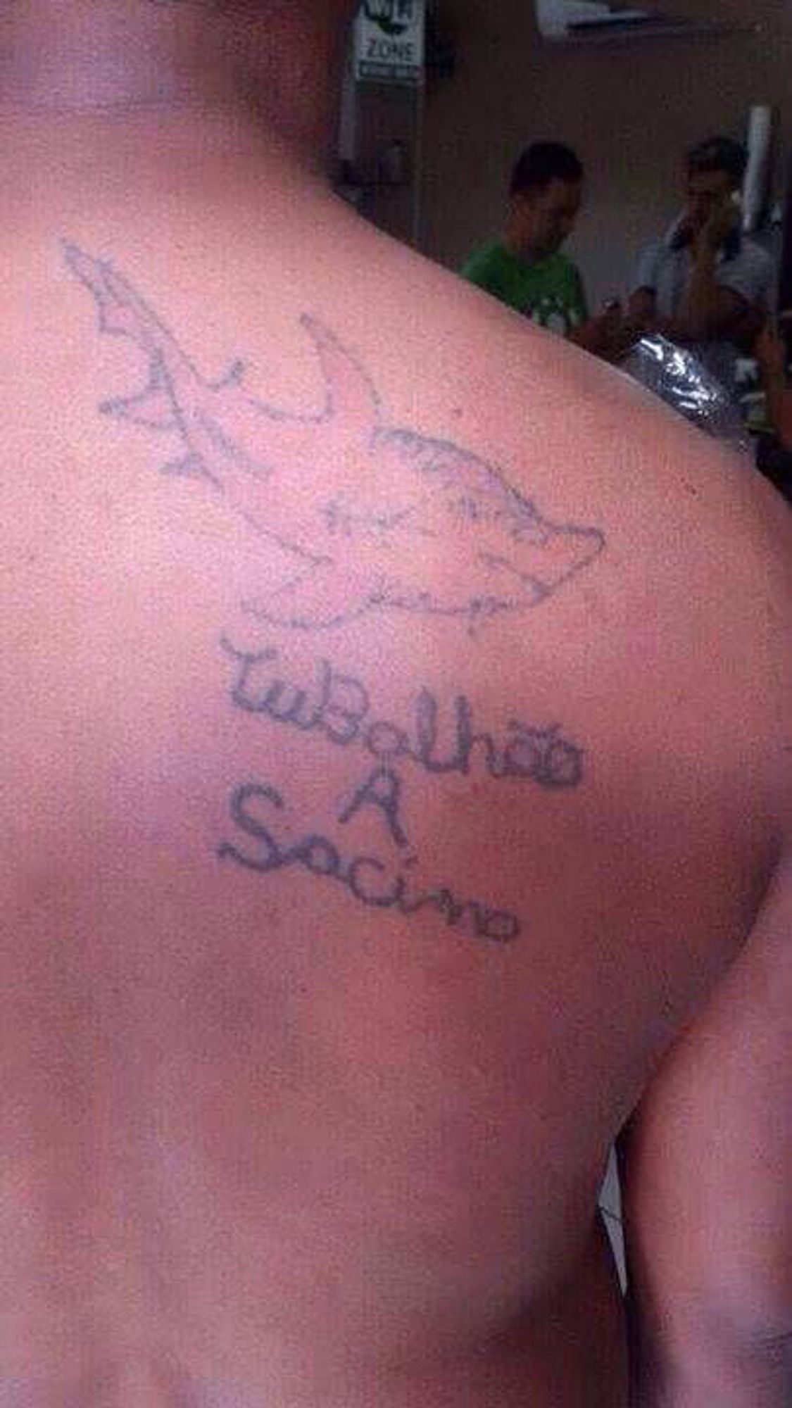 É uma foto de uma tatuagem nas costas de um homem consistente num desenho de tubarão muito feio e malfeito e embaixo está escrito tunalhão a sacino