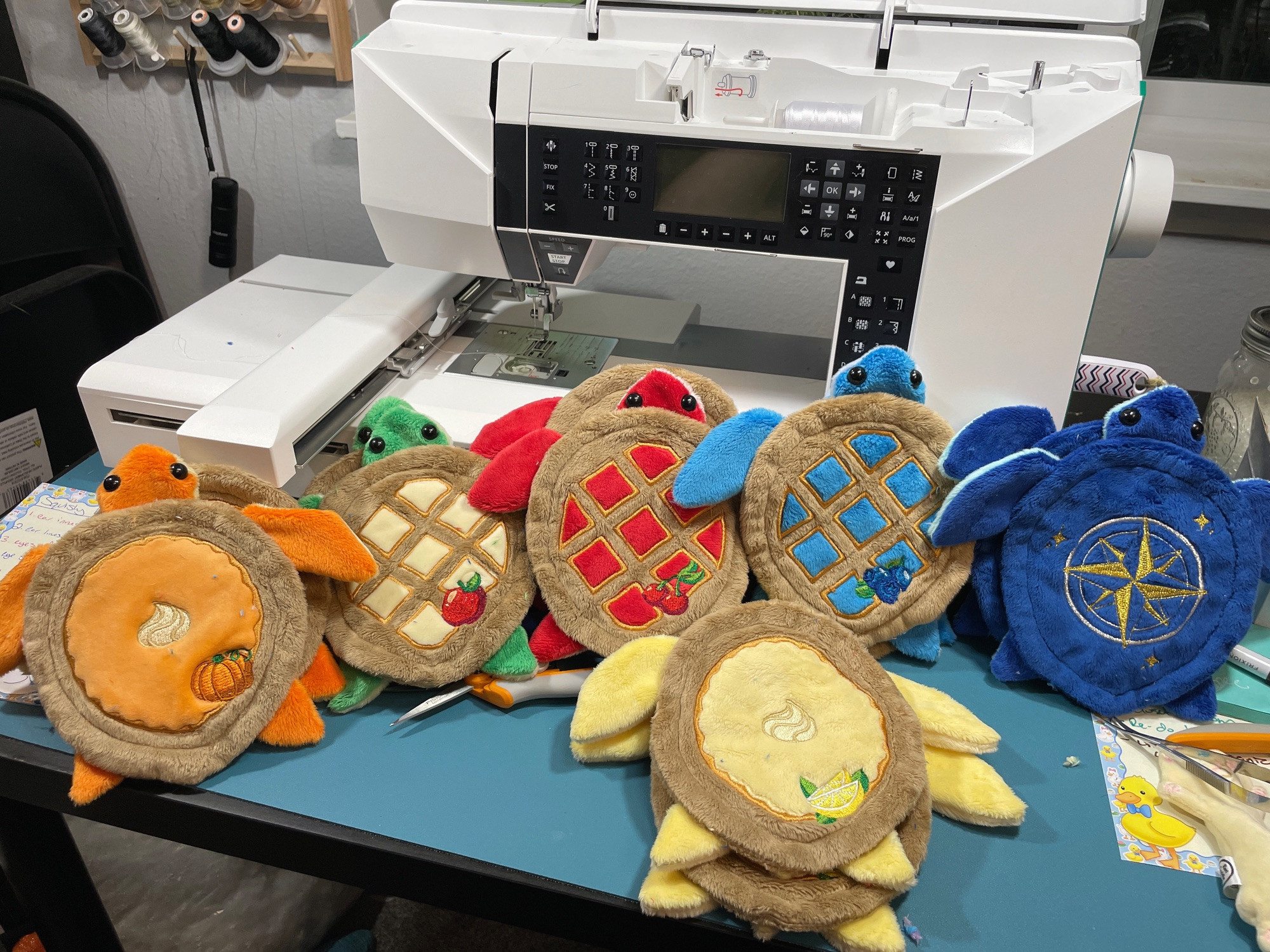 Many unstuffed turtle plushies on a desk with an embroidery machine in the background. Their shells look like pumpkin, apple, cherry, blueberry, and lemon merengue pie and one of them is blue with a gold compass on its shell.