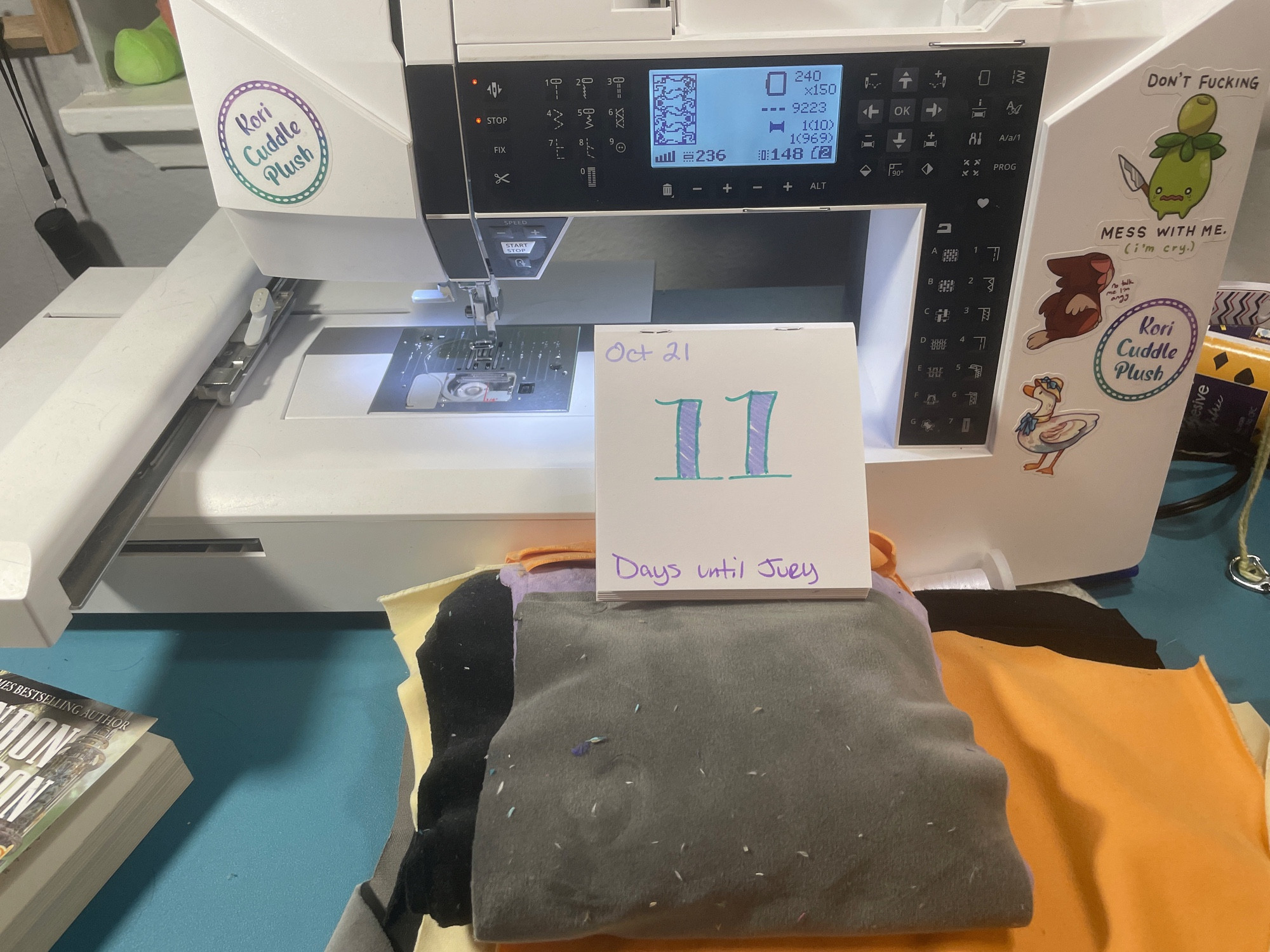 A photo of an embroidery machine and a stack of grey, orange, black, and cream fabrics. There’s a countdown calendar on top that reads “Oct 21: 11 days until jury”