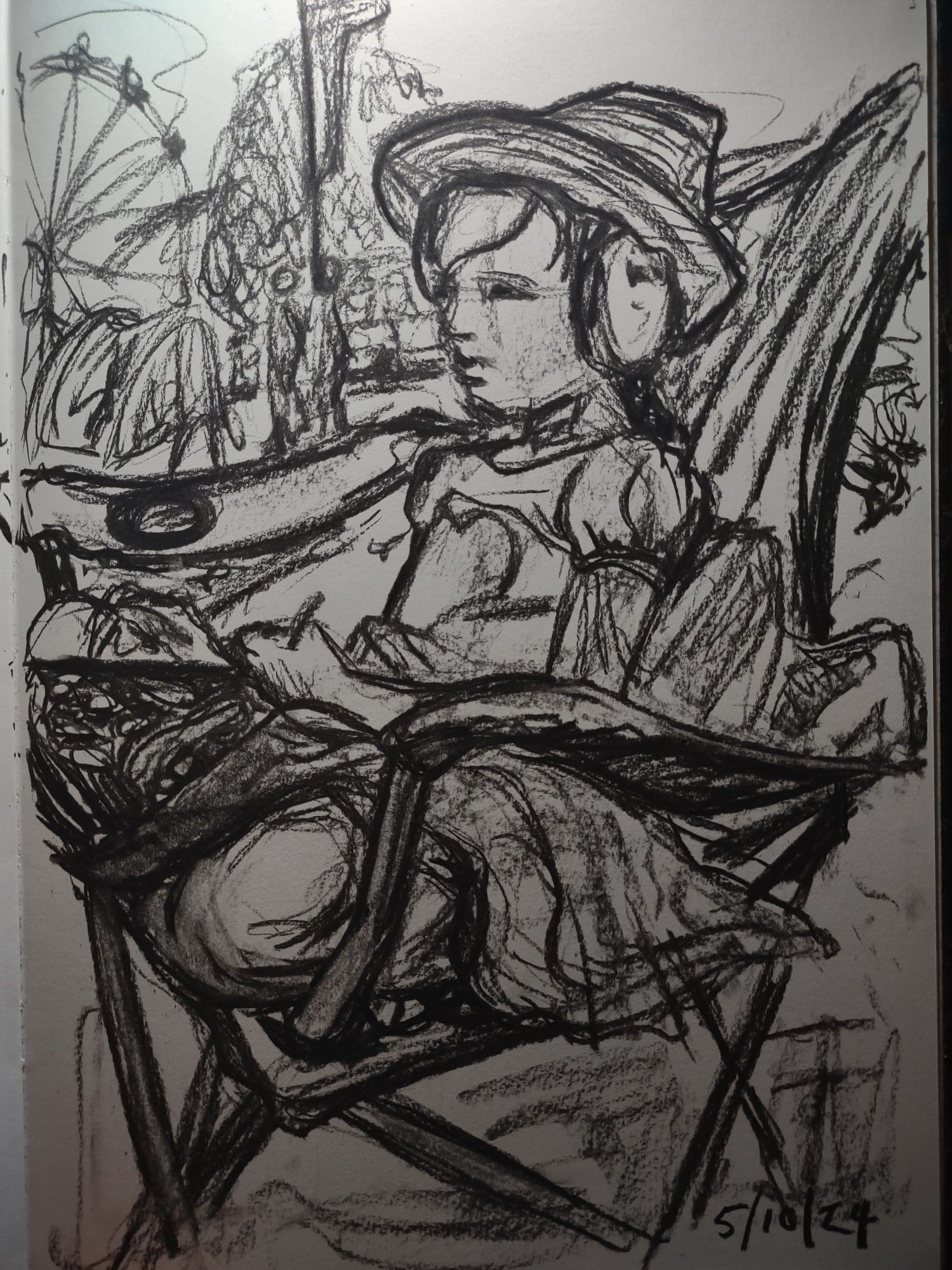 A charcoal sketch of a girl sitting cross-legged on a camping chair. She is facing left and looking into the distance as she draws on a pad of paper on her lap.