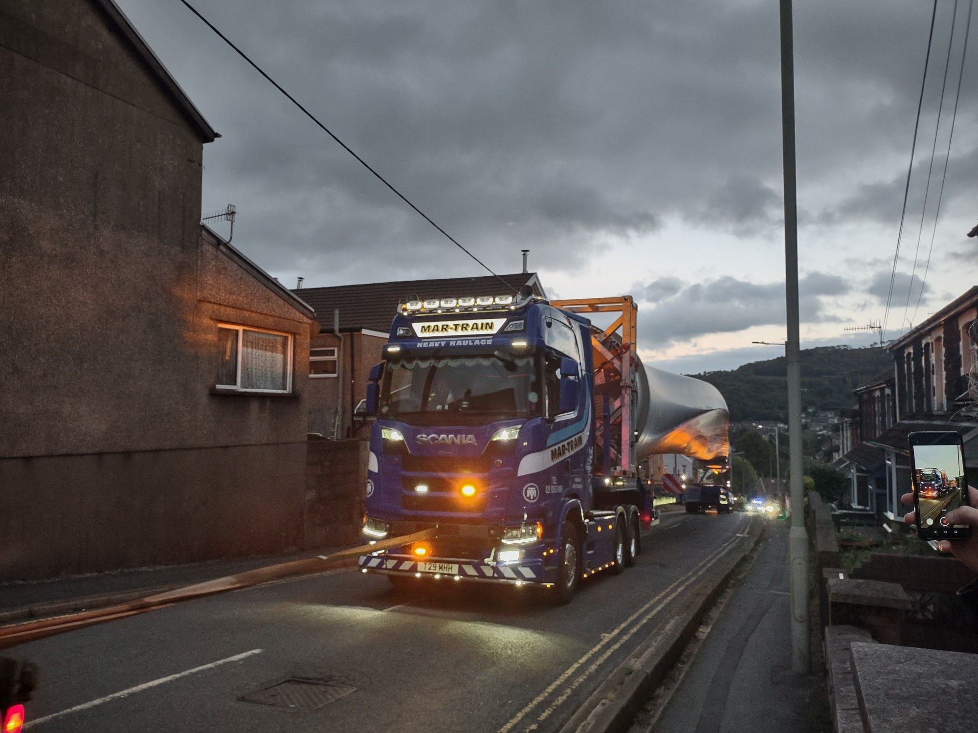 A photograph of a 55 m wind turbine blade being transported uphill along a residential street.