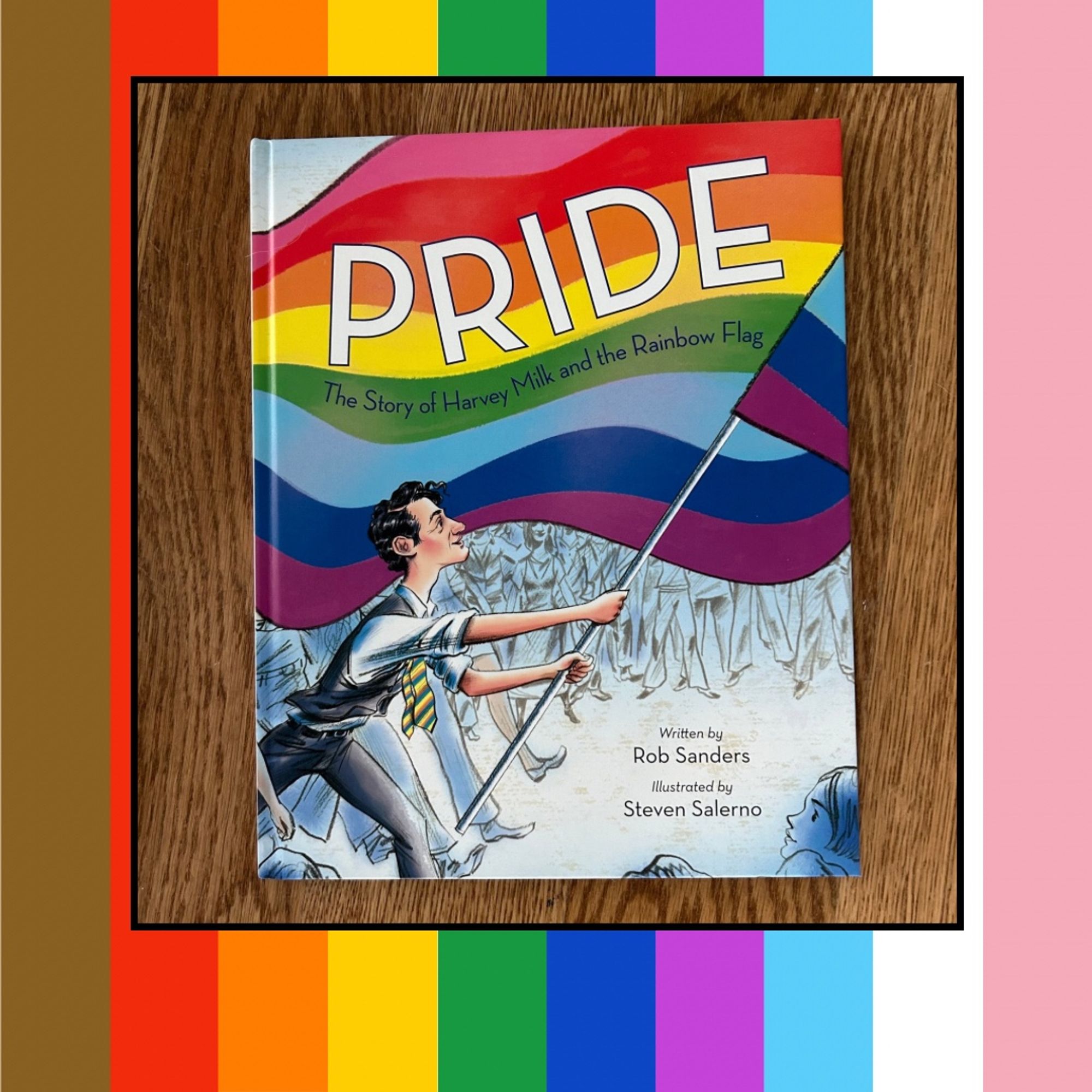 Rainbow striped graphic with a picture of a picture book on it. The picture book is Pride: The Story of Harvey Milk And The Rainbow Flag written by Rob Sanders and illustrated by Steven Salerno. The book cover shows a man holding a large rainbow flag and the title goes across the stripes of the flag.