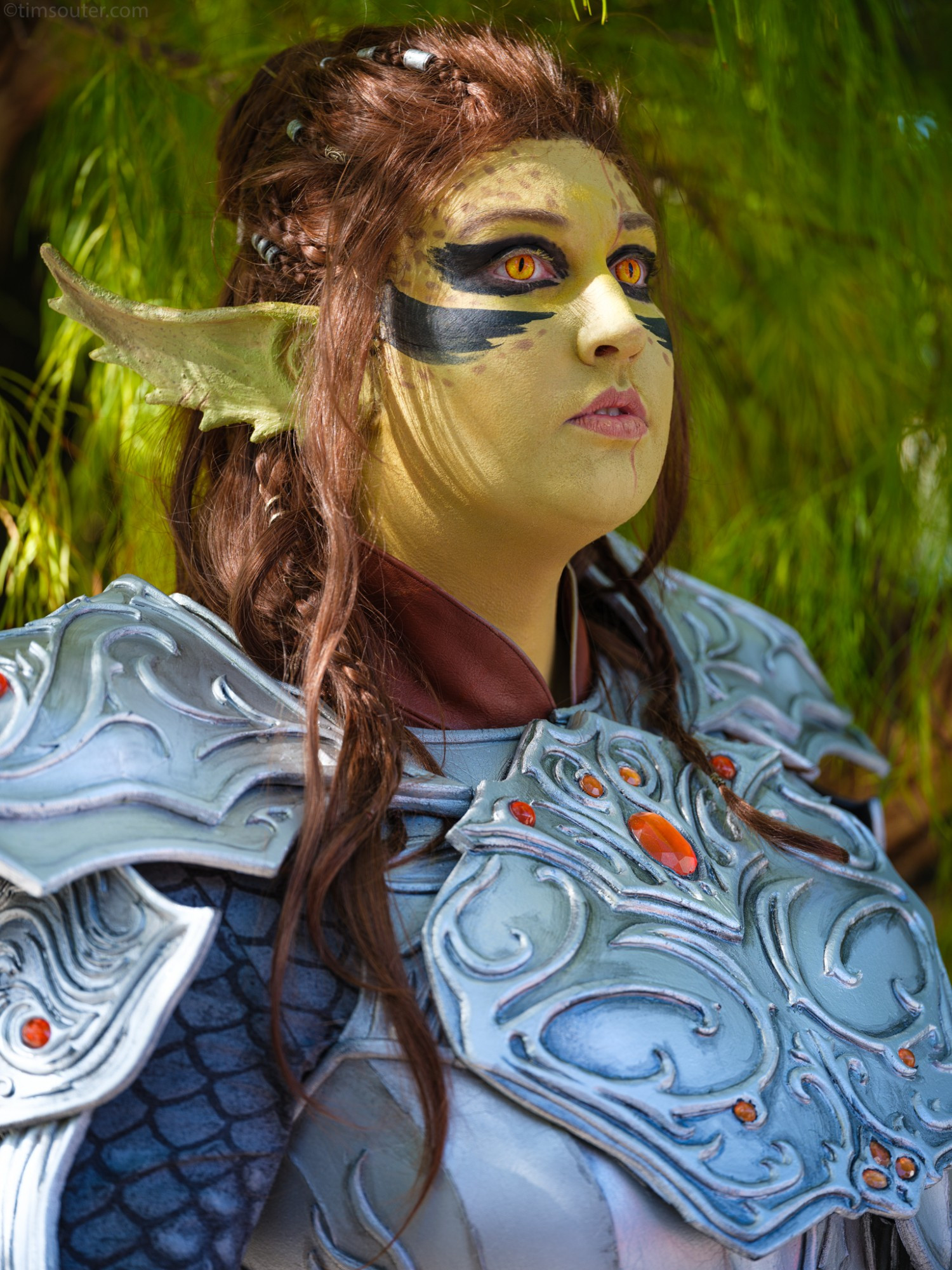 Scarlet Moth cosplays as Lae'zel from Baldur's Gate 3. The subject has greenish yellow skin, serrated elf ears, and brown hair done up in a bun with braids hanging loose. Her eyes, which are golden yellow with lizard-like slits, gaze away from the camera towards the right of the screen. She wears silver armour with ornate filigree and studded with red gems. Her face bears a neutral expression.