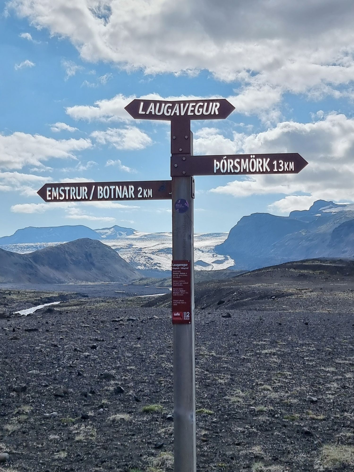 Laugavegur woodrn fingerpost against a background of rough black ground, with a glacier between distant peaks. The sign has the writing etch-cut out if it and the blue sky with puffy white clouds provides the 'font colour'! Left is Emstrur/Botnar 2km, and right Þórsmörk 13km. The top sign says Laugavegur.