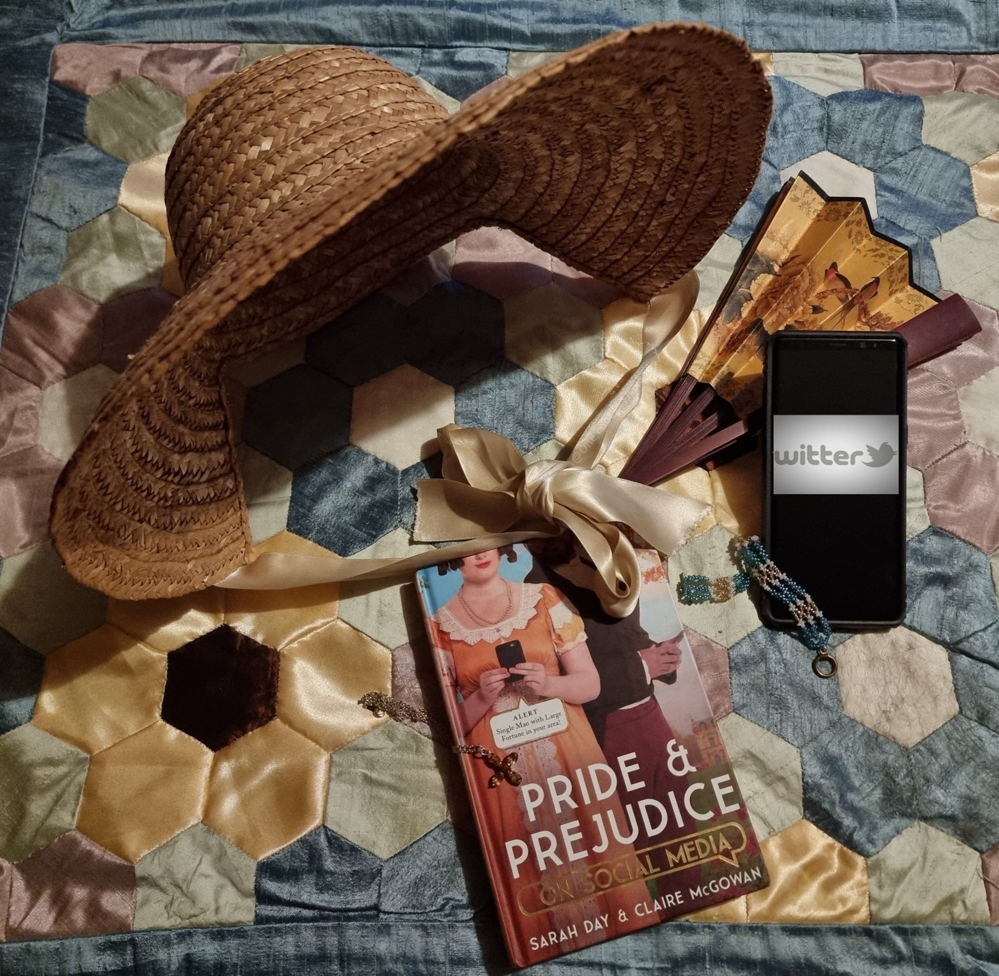 Straw bonnet, fan, phone, jewellery, and a copy of the book 'Pride & Prejudice on Social Media' by Sarah Day & Claire McGowan - the story told by its characters on twitter, group chats, Instagram etc - hilarious! All are laid on a background of a patchwork of hexagons in silks and satins of assorted colours 