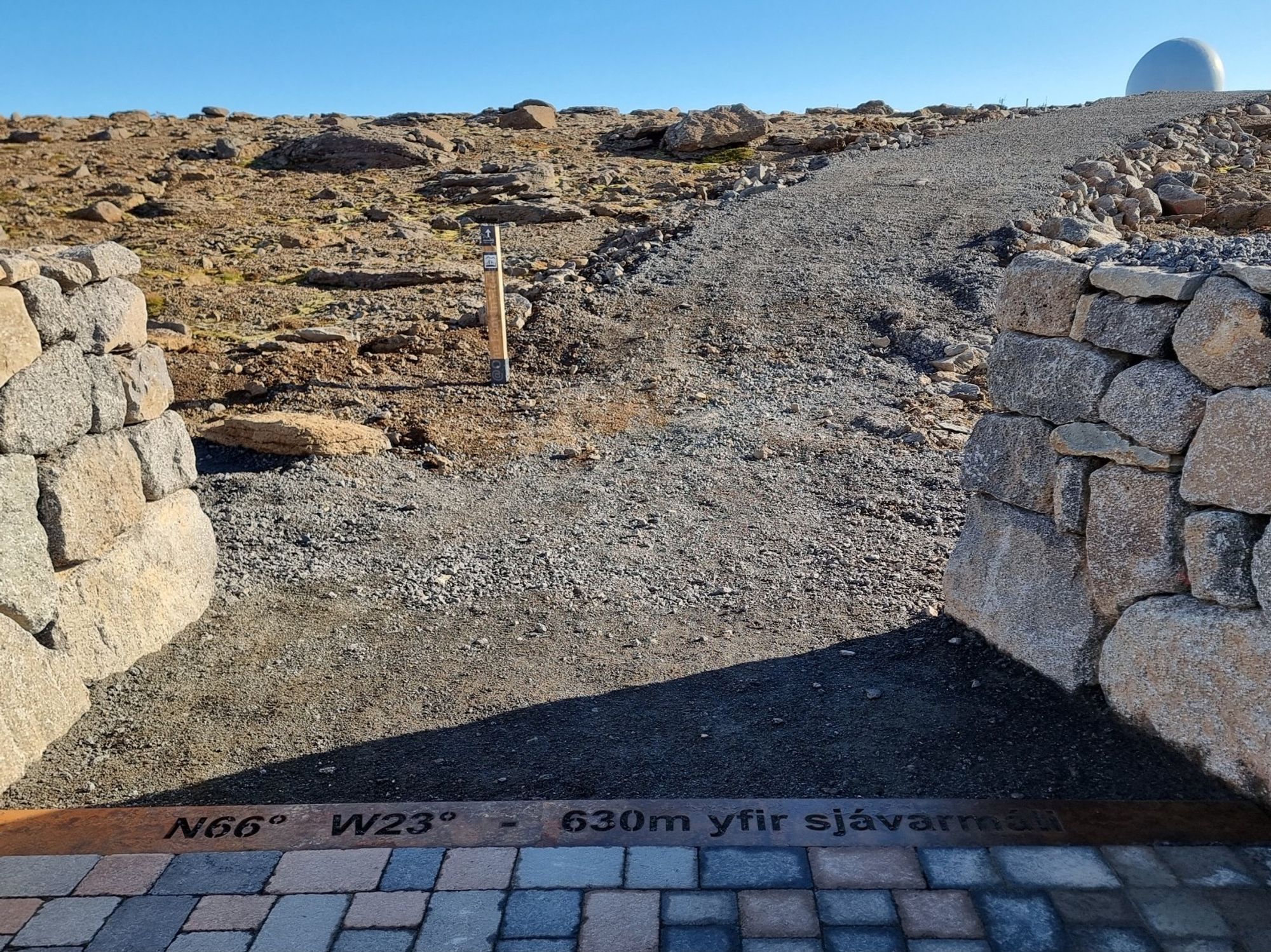 At the end of a paved path a strip on the ground says N66° W23° 630m over sealevel (yfir sjávarmáli). There are dry stone walls either side, and then a gravel path winds away between rough ground, with a globular radar tower visible on the horizon