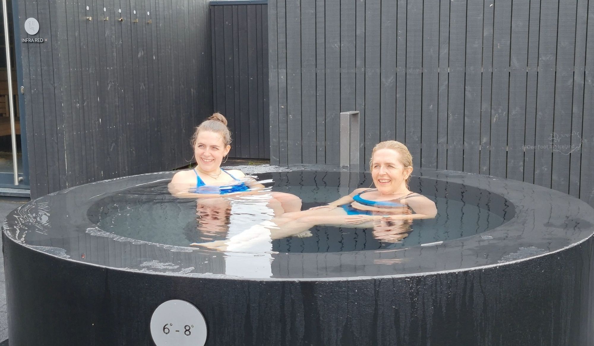 2 smiling white women in blue swimwear sit in a round black water pot with a sign showing the temperature to be 6-8°C. The walls behind are clad in black wood