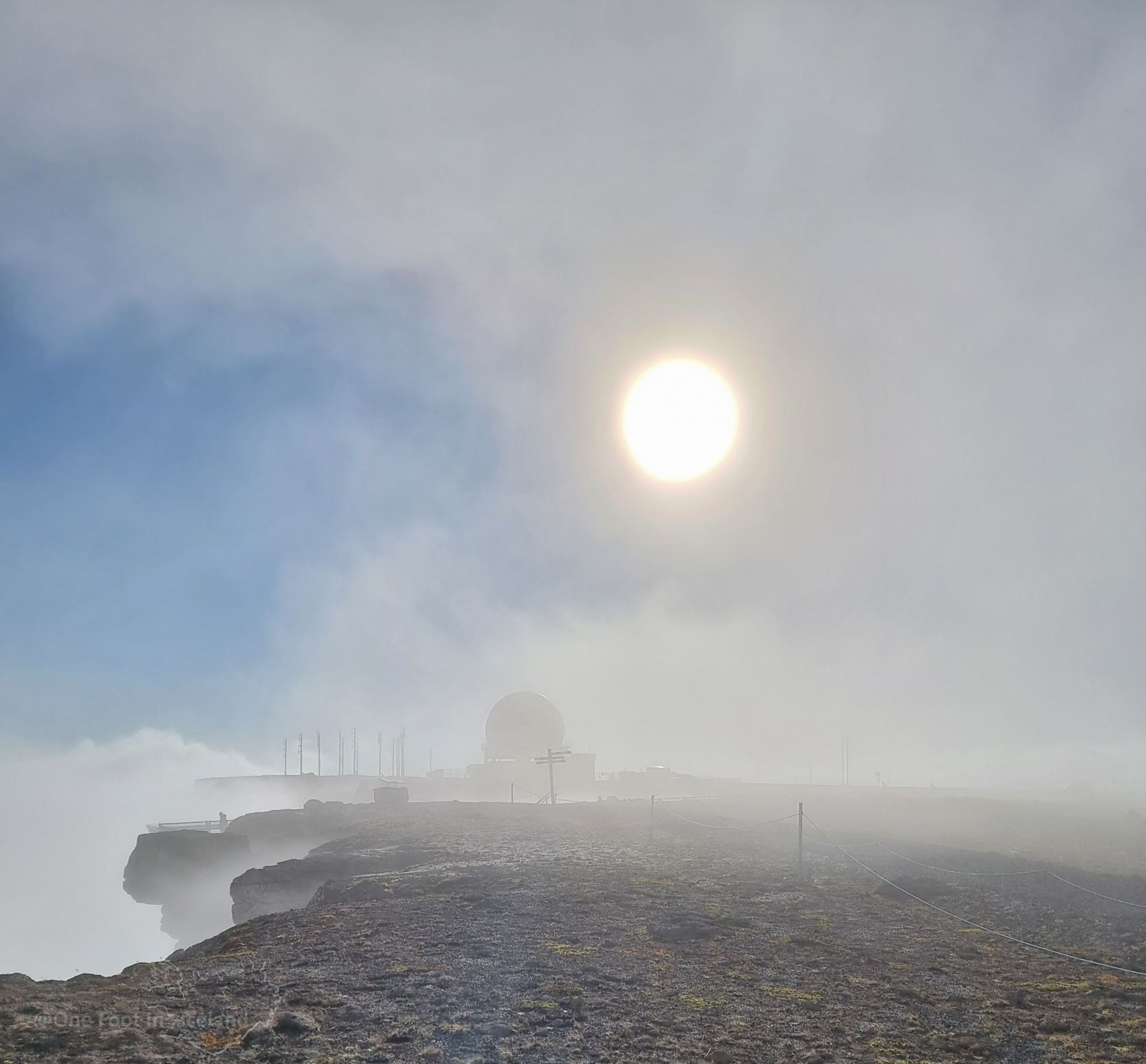 A stony cliff edge wreathed in mist, with a viewing balcony barely visible in the bottom left of the pic, with a man on it. The sun is shining through the mists and dimly reveals a radar station globular building and antennae