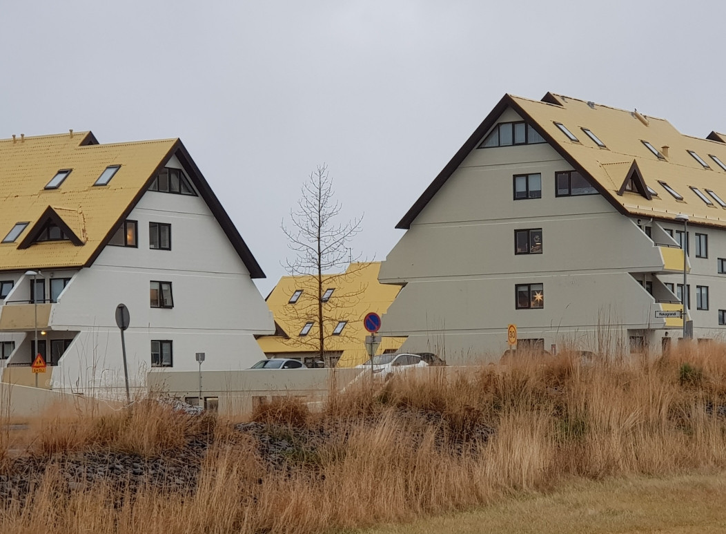 Two apartment blocks at right angles to each other, with yellowish roofs. The white end walls have zigzag edges giving the shape of Christmas trees 