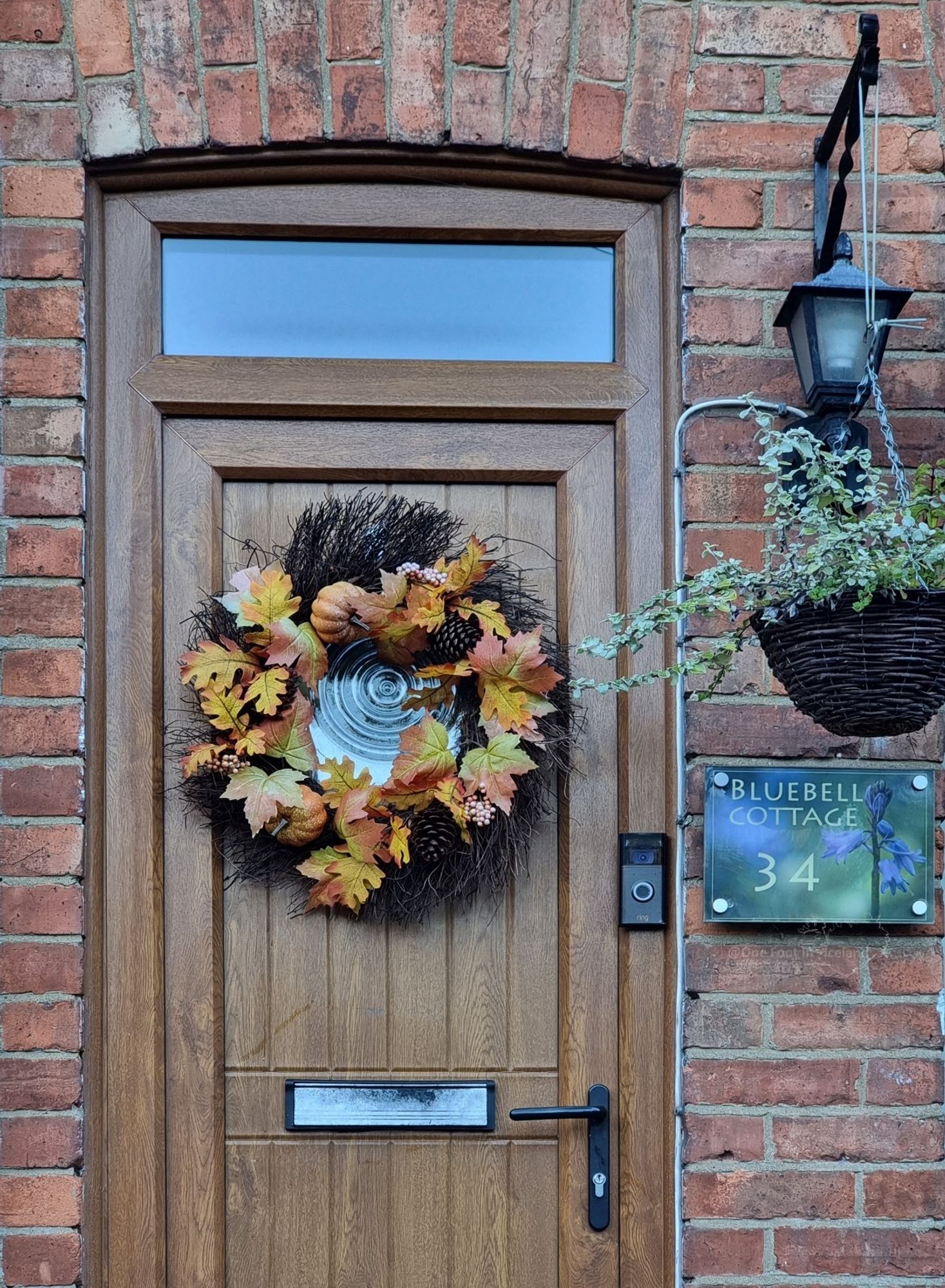 A mid brown wood effect UPVC front door with a twig wreath decorated with autumn leaves hanging around a circular window of rippled glass. There is black door furniture, and a hanging basket on the right with a plant in, and carriage lamp above, and a sign for the house 'Bluebell Cottage' No. 34