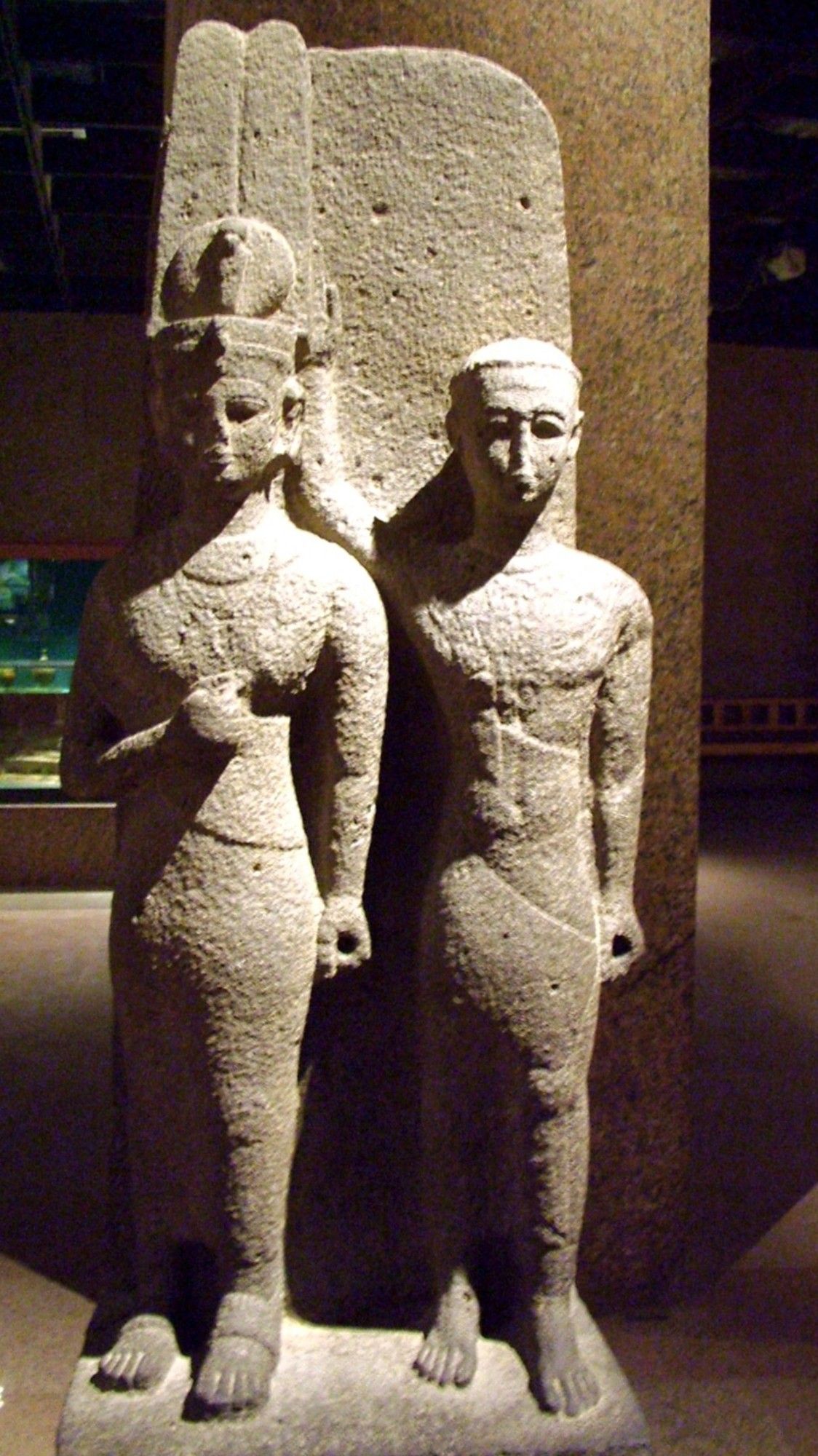 double stone statue of Nilotic african queen and male companion. her right arm is raised, resting above her stomach. the other's arms are down to his sides.