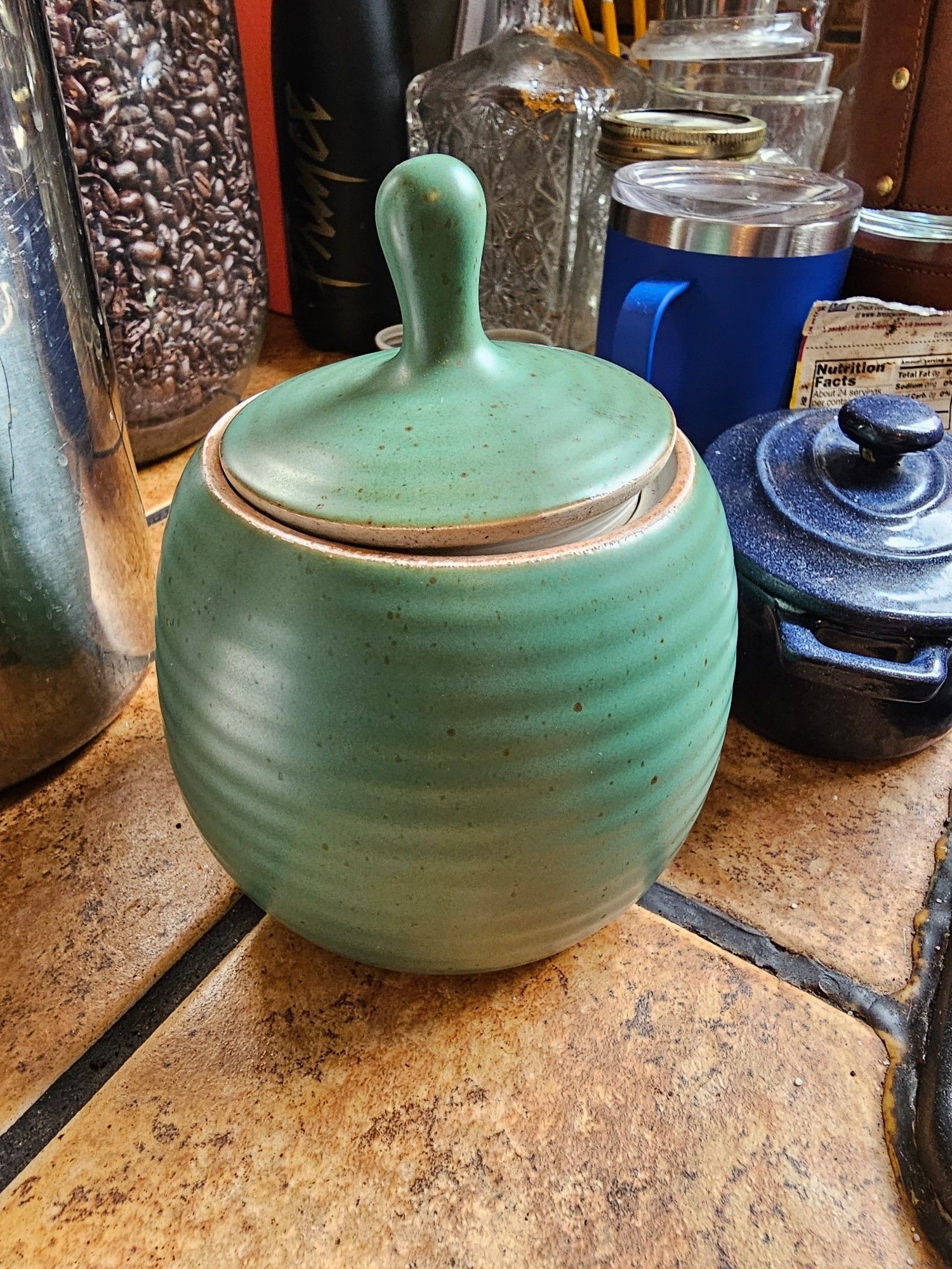 green ceramic pot with pointy-handled top. in the background are a blue dutch-oven styled butter dish and a large glass jar of wholebean coffee.