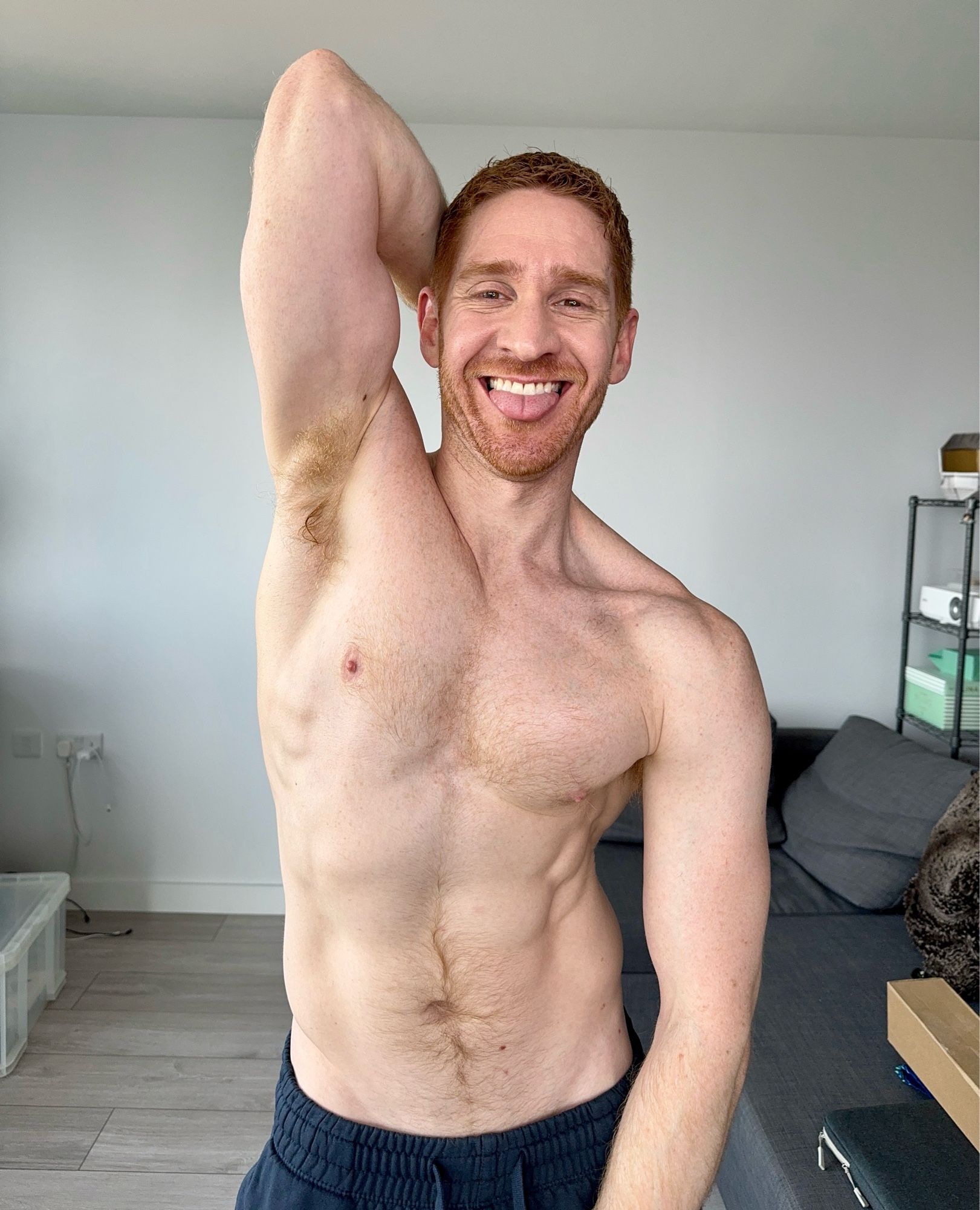 Leander, a white redhead, stands topless in navy sweatpants. One arm is behind his head exposing his ginger armpit. He has a big smile and is poking his tongue out