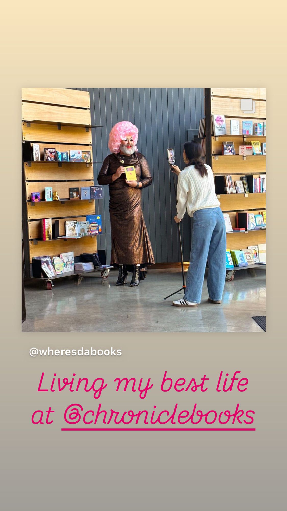 The image shows a person in a pink wig, dressed in a metallic gown, holding a book while being photographed or recorded by another individual with a smartphone on a tripod. They are standing inside what appears to be a bookstore with shelves full of books behind them. The text on the image reads: "Living my best life at @chroniclebooks" and includes the Instagram handle "@wheresdabooks."