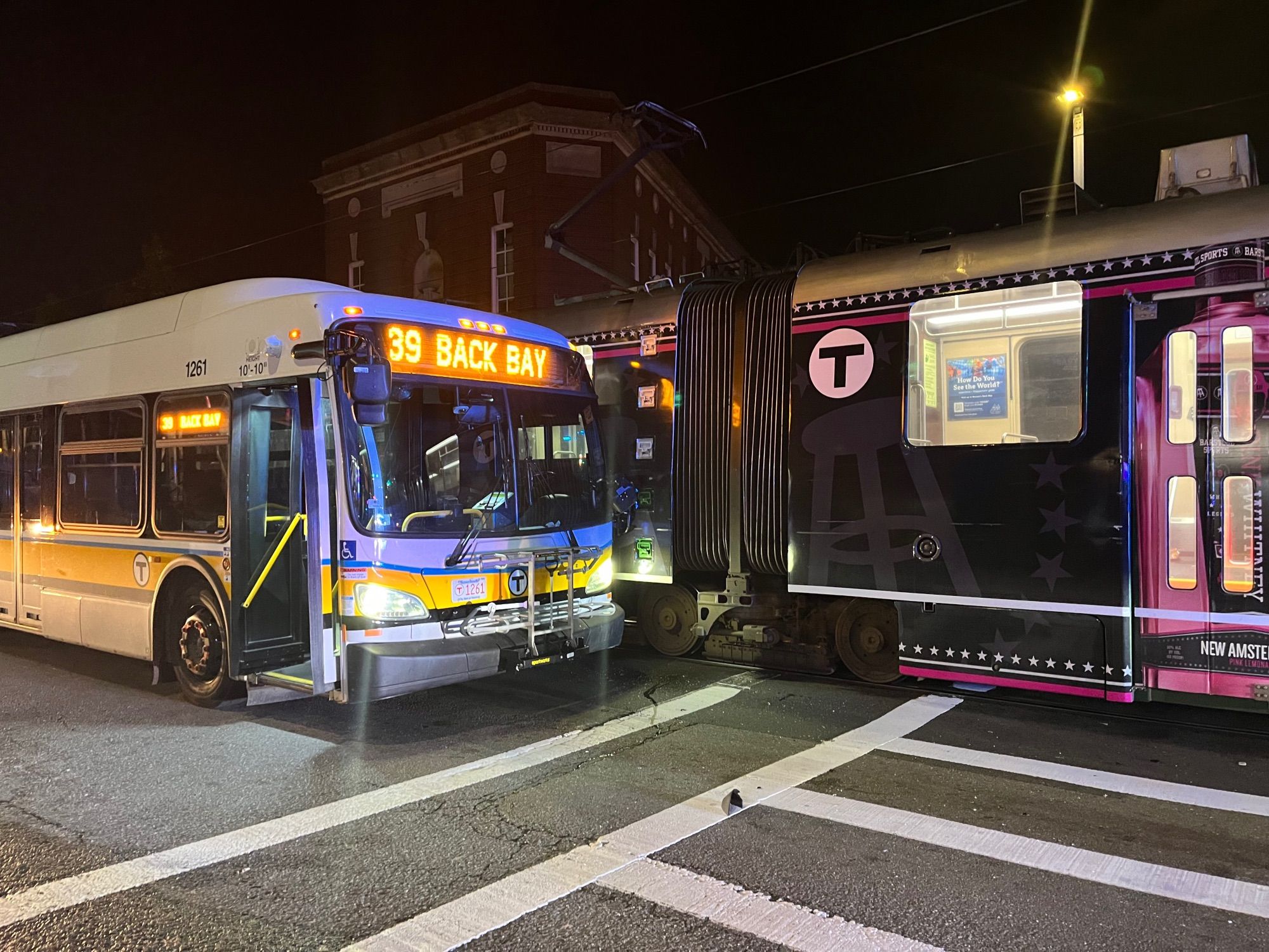 At a crosswalk, a 39 Back Bay bus has made contact with a Green line train. A streak along the train body indicates initial contact may have been further forward on the train.