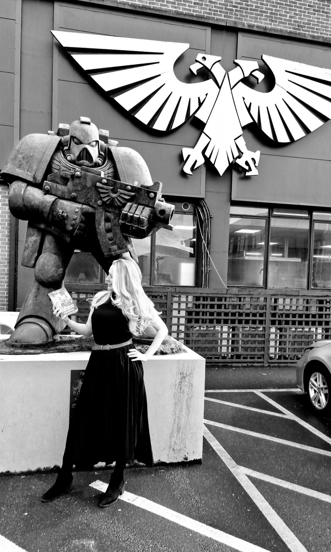 A black and white photo of Victoria holding a copy of Deathworlder in front of the Games Workshop car park Space Marine