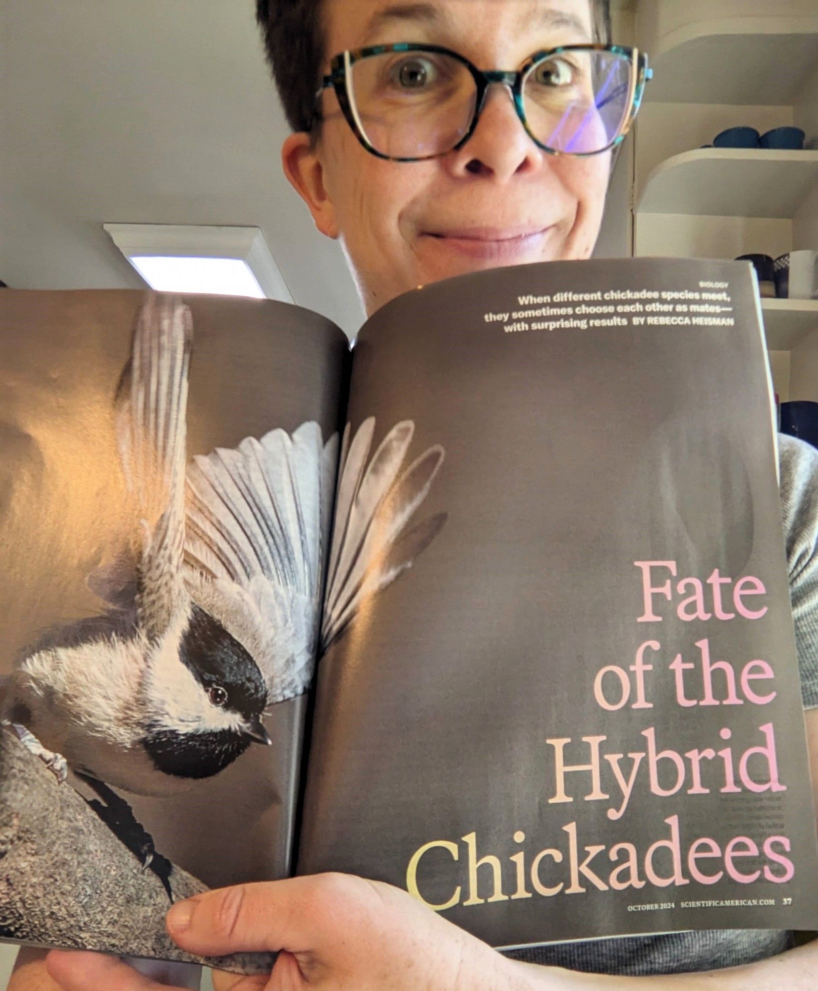 an excited-looking woman with short hair and glasses poses with a magazine, open to a spread with a large photo of a chickadee and the large text "Fate of the Hybrid Chickadees"