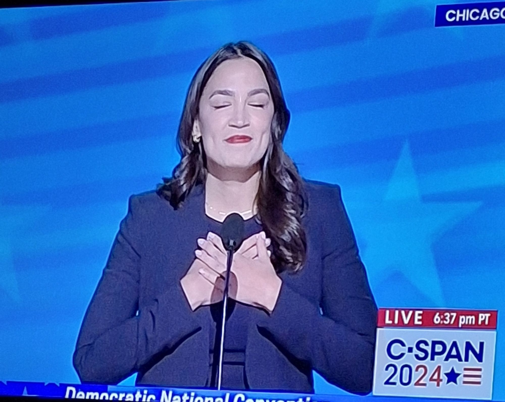 AOC with eyes closed, hands crossed over her chest, smiling at the start of her DNC speech while the crowd is chanting her initials.