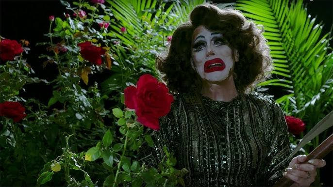 Sharon Needles in a rose field wearing a clownesque white make up with deep red lipstick, a short Joan Crawford style wig and carrying a battle-axe 