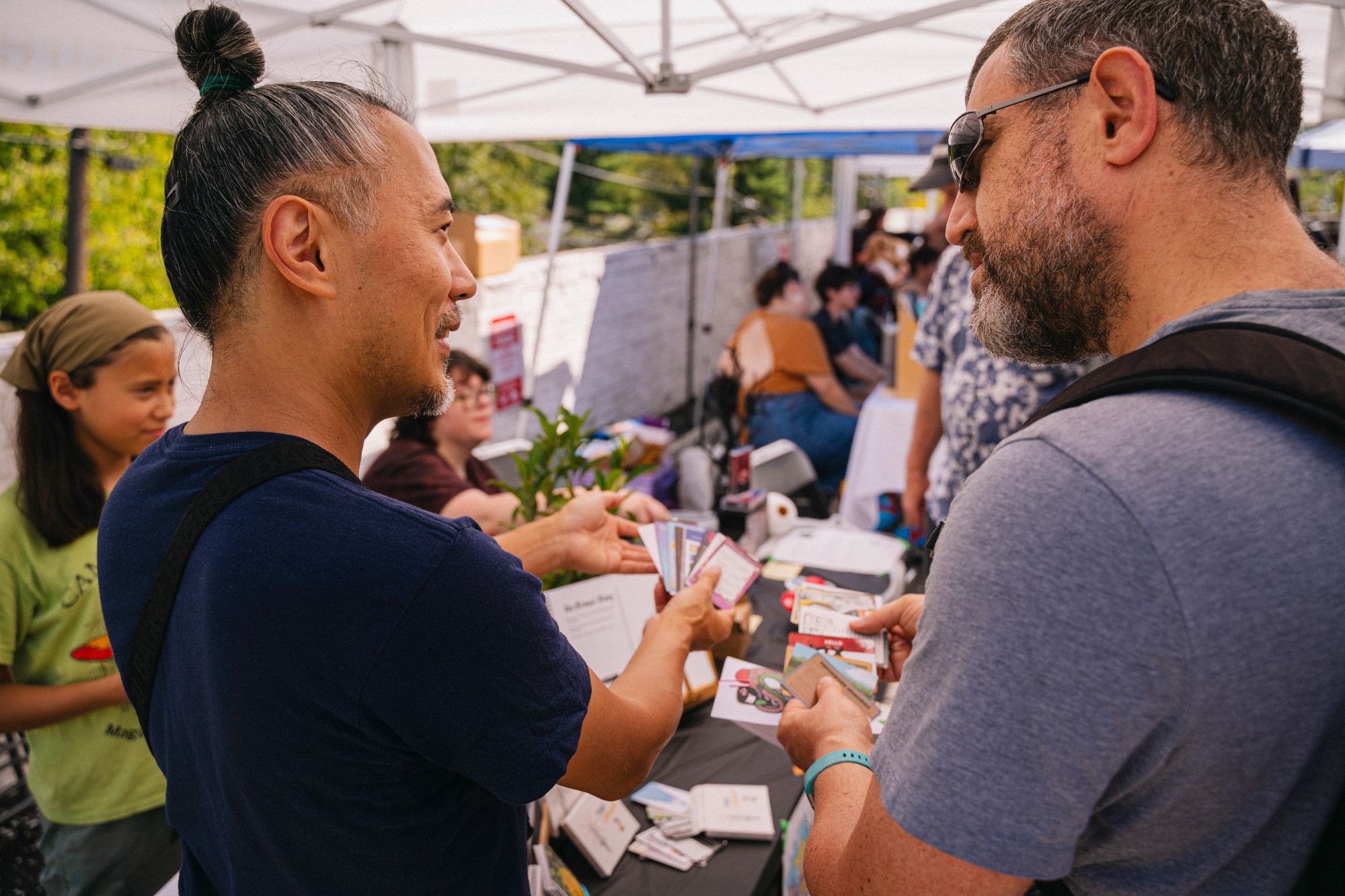 Gene Koo (Sticky Doodler) showing cards from the Tiny Library series to a potential customer. The setting is outdoors, with tents covering tables.