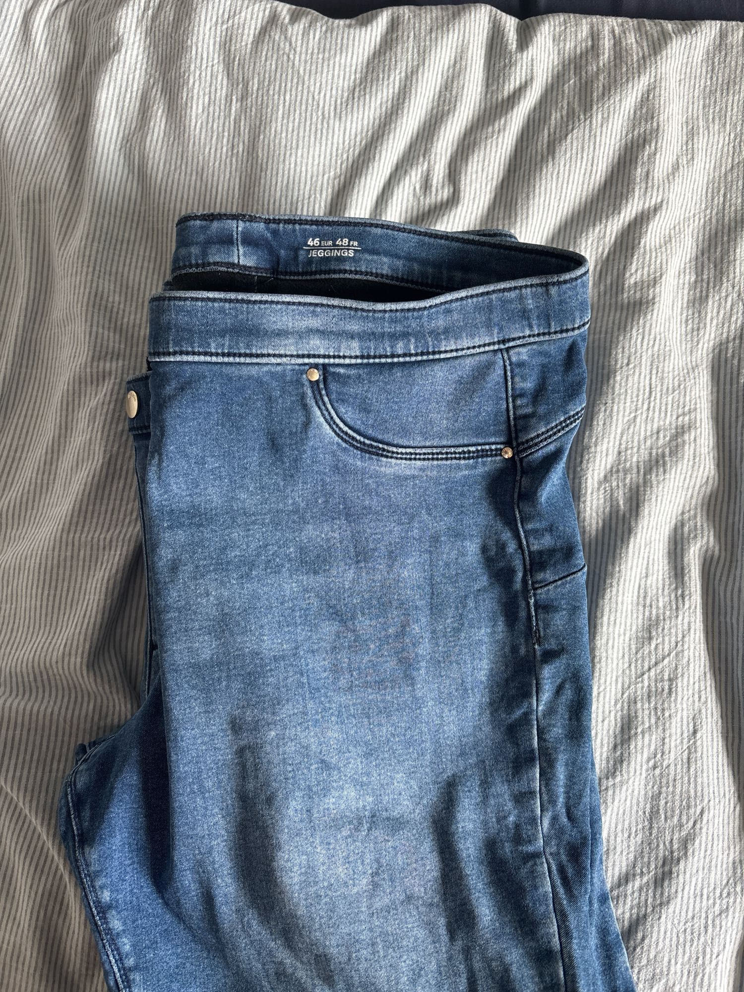 Man sieht die obere Hälfte einer gefalteten blauen Jeans auf einem hellblau-weiß gestreiften Untergrund. Im Bund der Jeans steht geschrieben: 46 EUR, 48 FR, Jeggings