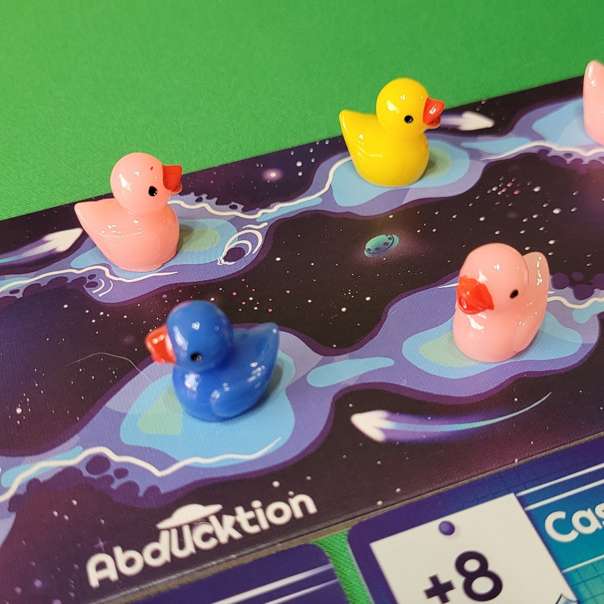 Pic of part of Abducktion board showing 4 ducks in the galactic pond, lined up to be "abducked". Pink and yellow on the top row, blue and pink on the bottom. The small arrows on the board show the direction the ducks move when filling in gaps after being abducted.