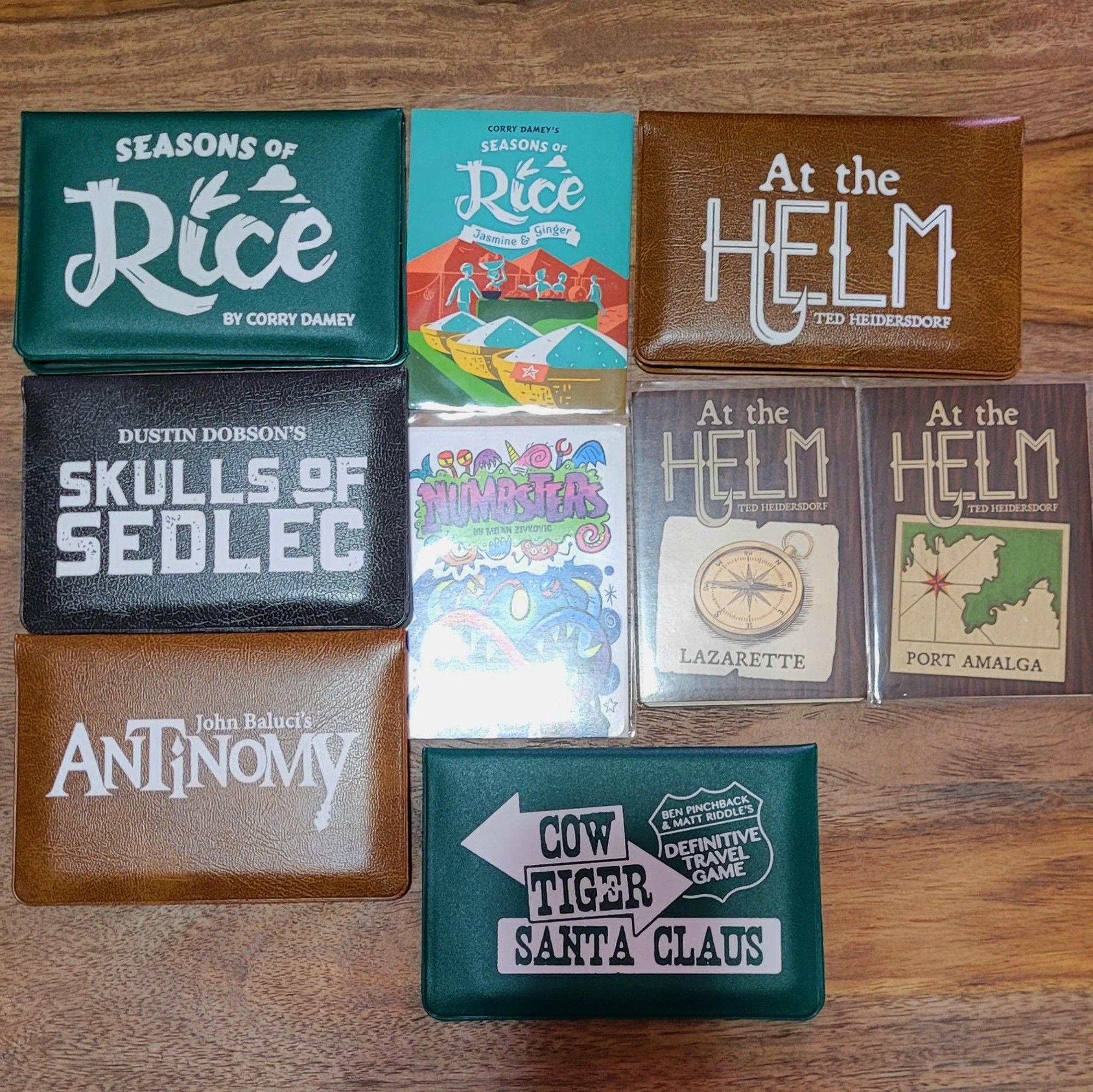 Wallet games, with some expansions in clear sleeves. Left column, top to bottom: Seasons of Rice (green wallet), Skulls of Sedlec (black wallet), Antinomy (brown wallet) . In the middle, top to bottom: Seasons of Rice Jasmine and Ginger expansion (clear sleeve), Numbsters Infinity (clear sleeve, 2 cards) expansion. Bottom center: Cow, Tiger, Santa Claus (green wallet, a long out-of-print travel game). On the right, at top: At the Helm (brown wallet) . Below it, 2 expansions in clear sleeves: Lafayette, and Port Amalga.