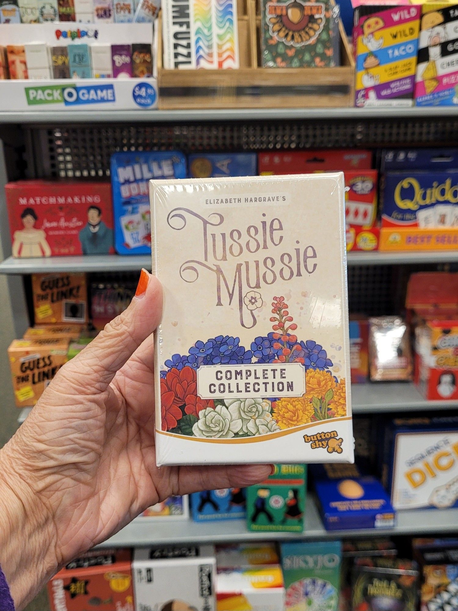 Holding up the small box of Tussie Mussie Complete Collection. It'll almost fit in my hand! In a Barnes and Noble store. Backdrop is a rack of very small box games.