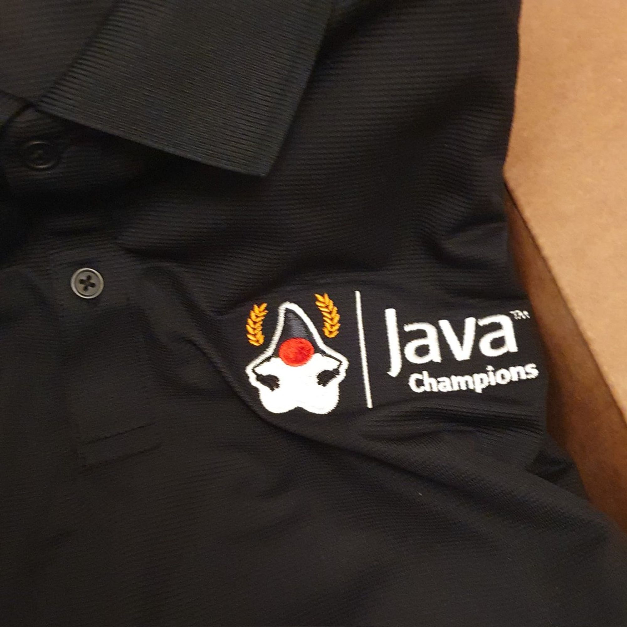 A black polo with the Java Champions logo sitting in a cardboard box