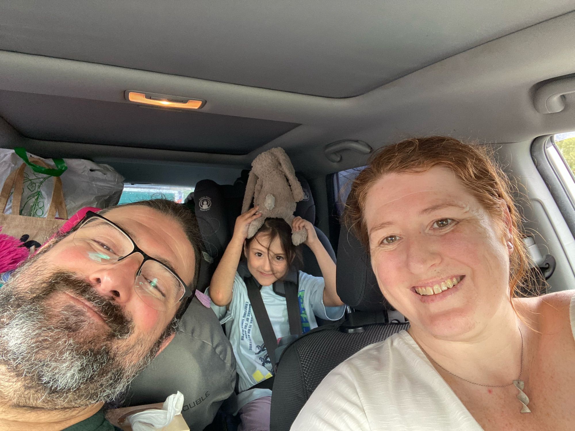 Picture of me, my Maltese partner and our 6 year old daughter packed into a very full car for a camping trip. Everyone is smiling and happy.