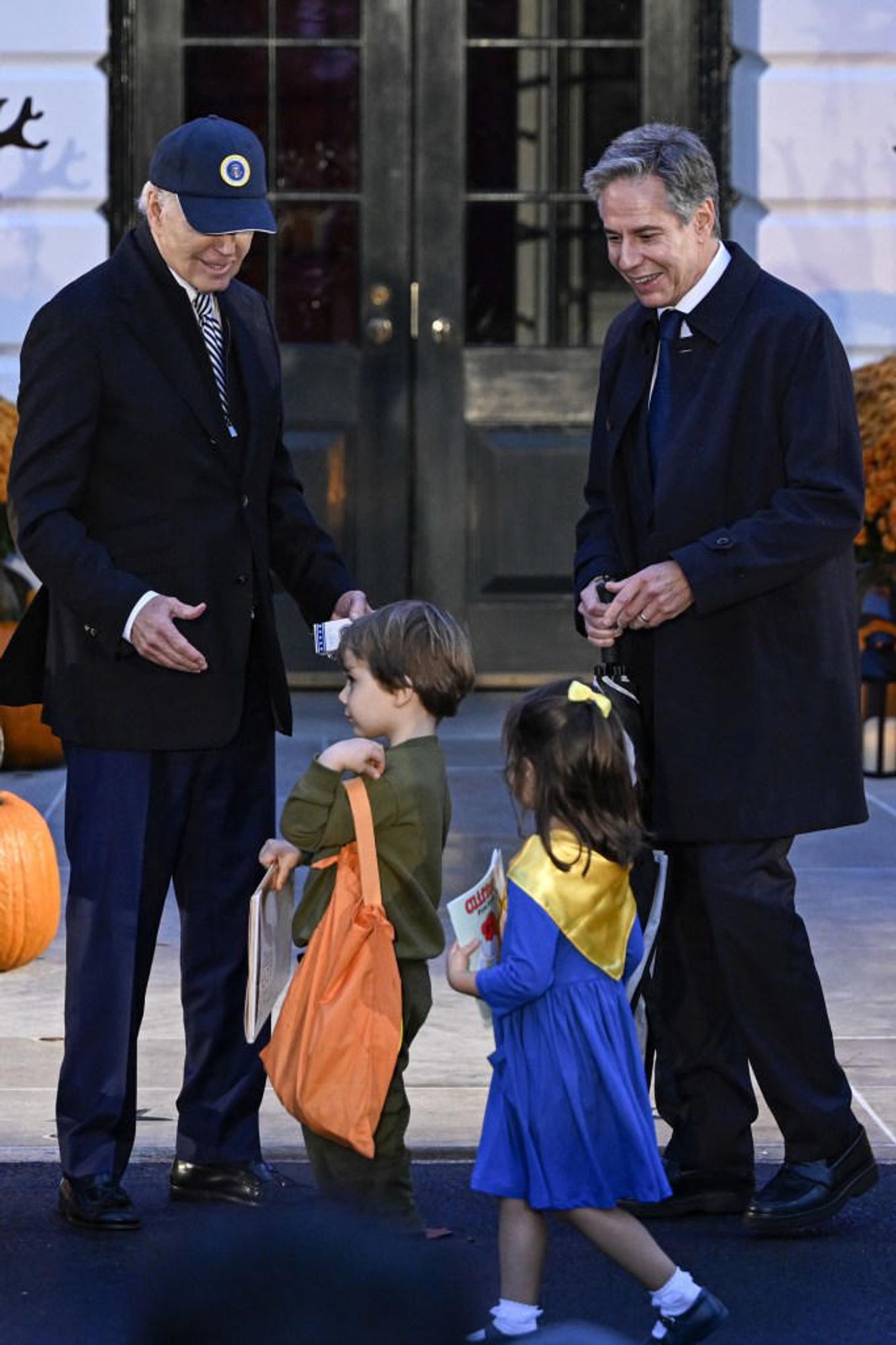 Biden smiling down at Blinken's kids: one is wearing an army green top and pants, the other is wearing a blue and yellow dress with a yellow bow in their hair. Blinken is smiling as he guides them over to Biden.