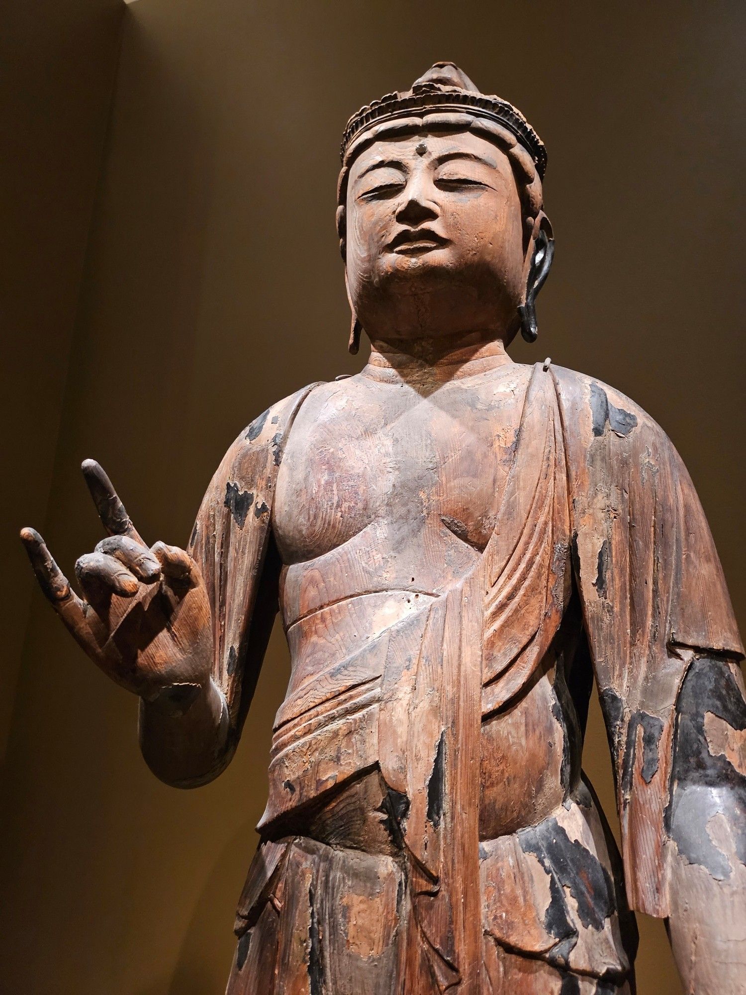 A carved wood buddhist statue from the san francisco asian art museum, making the sign of peace with its right hand, which kinda makes it look like it is throwing up metal horns.