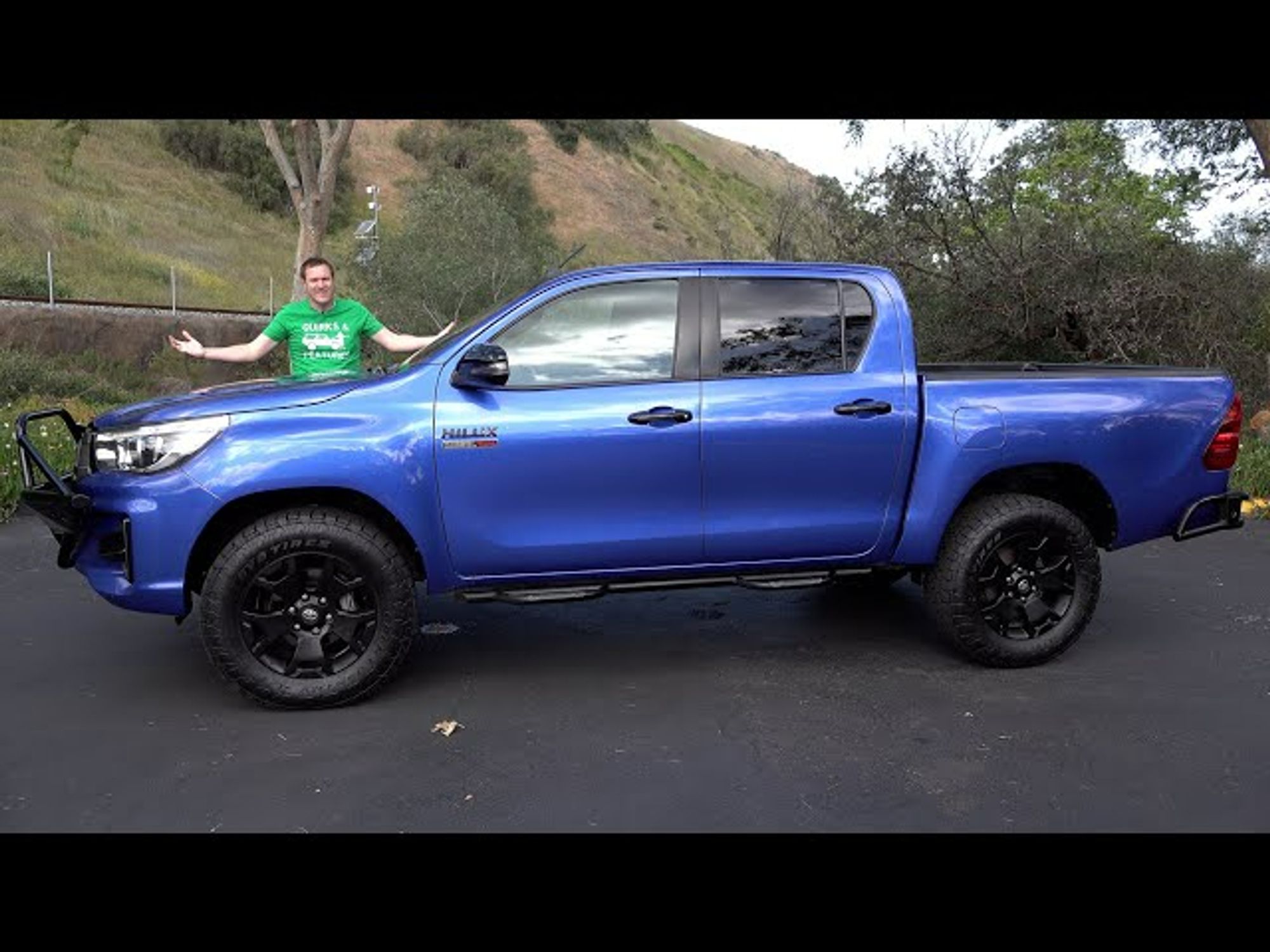 Doug Demuro making his trademark preview image pose in front of a Toyota Hilux, whose low, downward-sloping hood is more obvious in this pic. At its highest point, by the base of the windshield, it only barely appears higher than his waistline