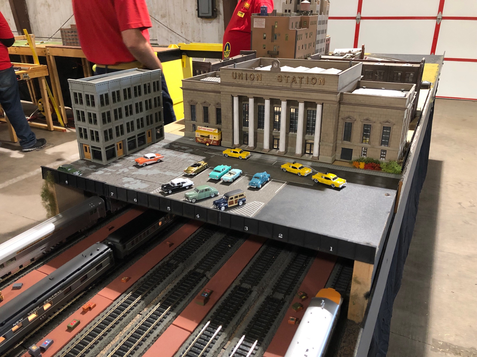 Photo of an HO scale module with multiple tracks disappearing under a roadway bridge with the Walthers Union Station kit poised above them, an Art Deco bastion of rail travel