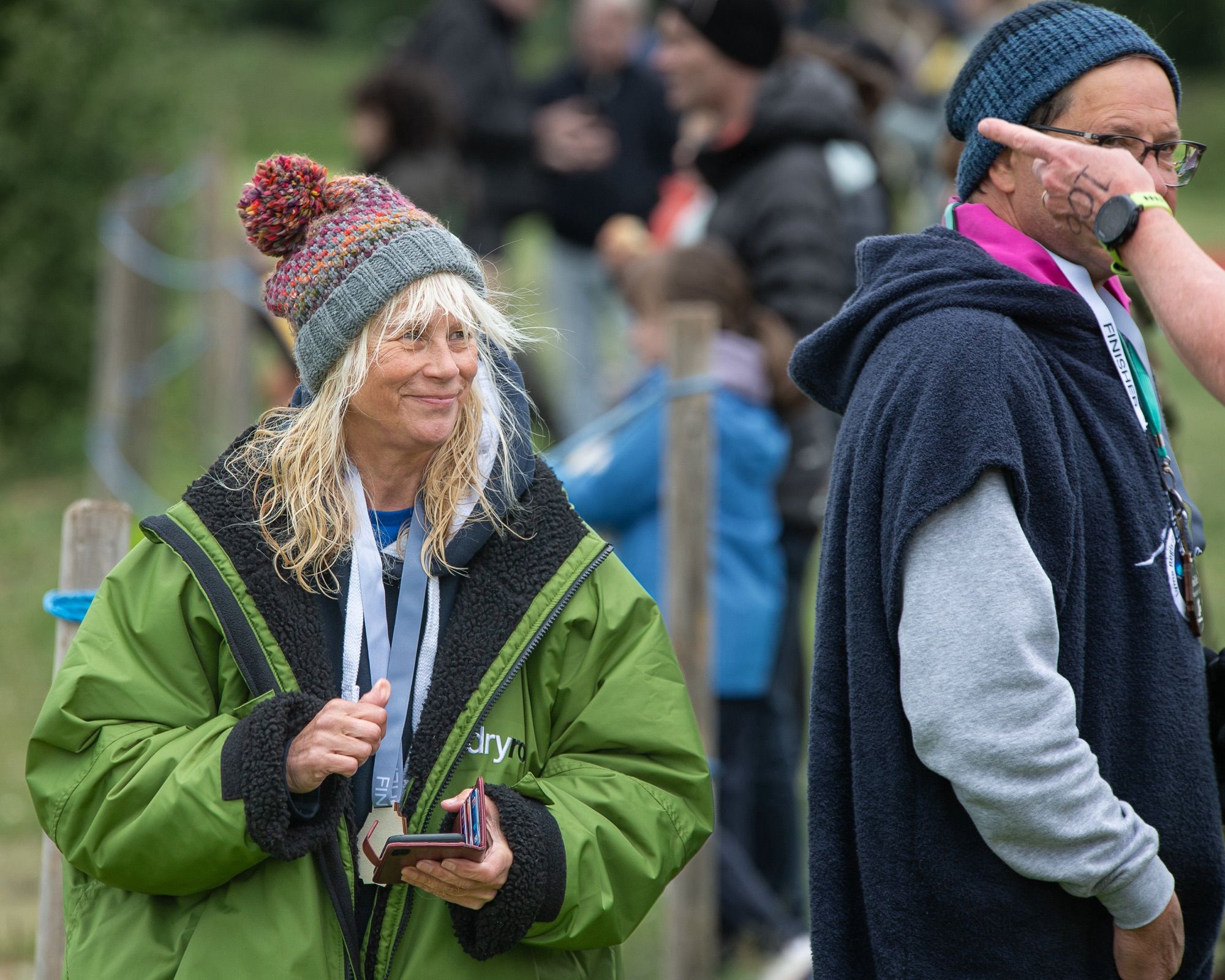 A swimmer who has completed their open water swim in a woolly hat and DryRobe holding mobile phone and smiling