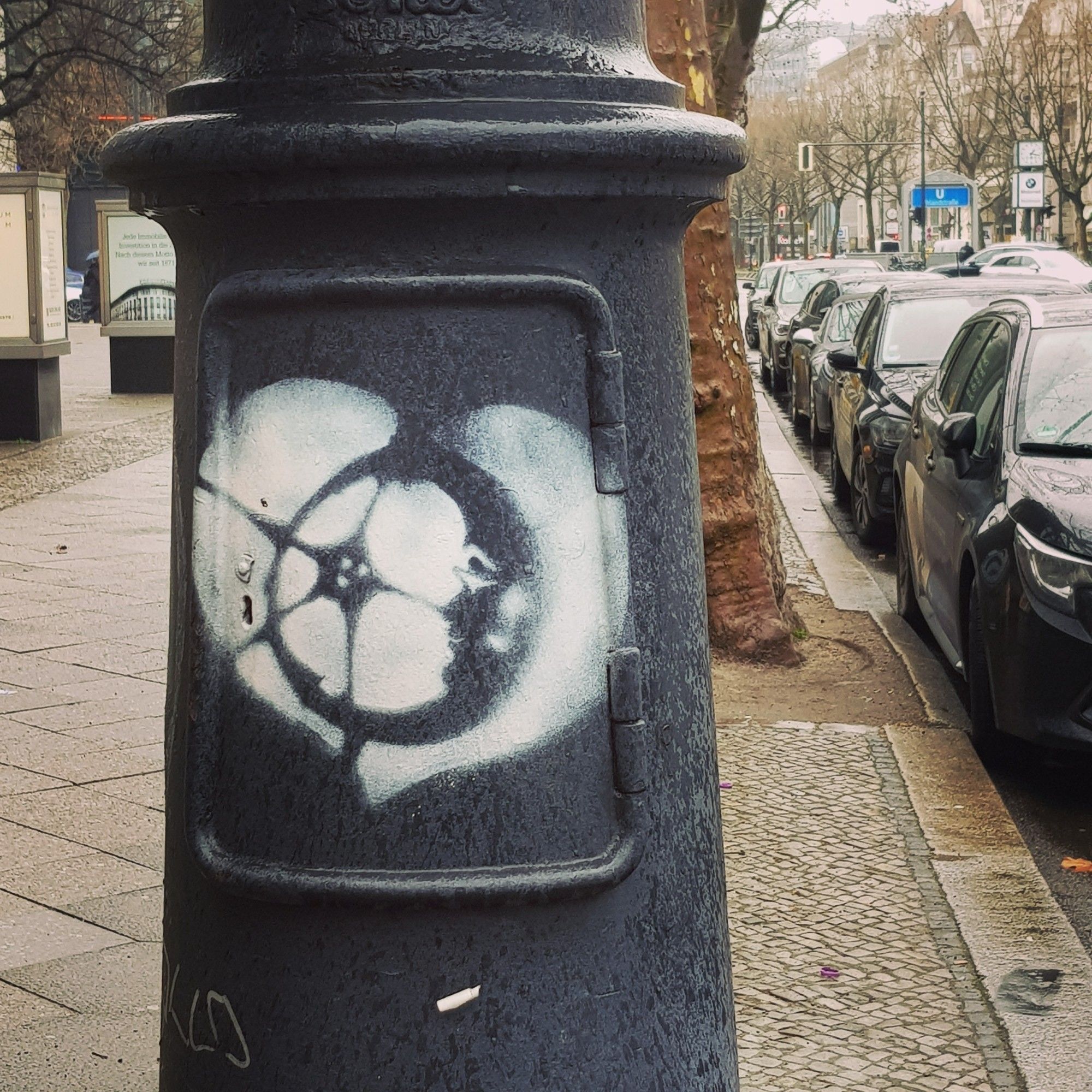 Blick die Straße entlang: ein gepflasterter Bürgersteig, rechts gesäumt von parkenden Autos. Der Blick/die Kamera fokussiert den Sockel einer alten Straßenlaterne, auf dem ein Graffiti in weißer Farbe (mit Schablone aufgebracht) prangt: ein Herz, inmitten die Mondsichel mit freundlichem Gesicht im Profil.

View along the street: a cobbled sidewalk, lined with parked cars on the right. The view/camera focuses on a graffiti in white paint (applied with a stencil), emblazoned on the base of an old street lamp: a heart, in the middle the crescent moon with a friendly face in profile.