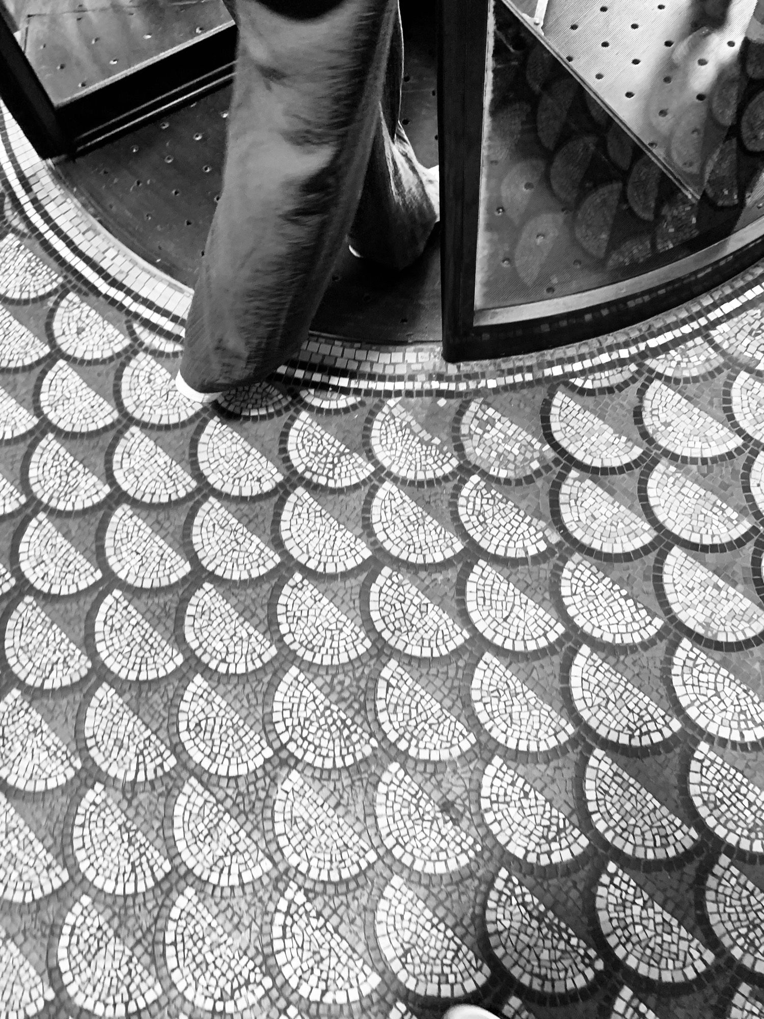 Picture of a mosaic doorway, there are semi circles of mosaic tiles plus a semi circle of the revolving door.  Somebody is stepping into the revolving doorway.  It’s busy and yet so restful to look at.