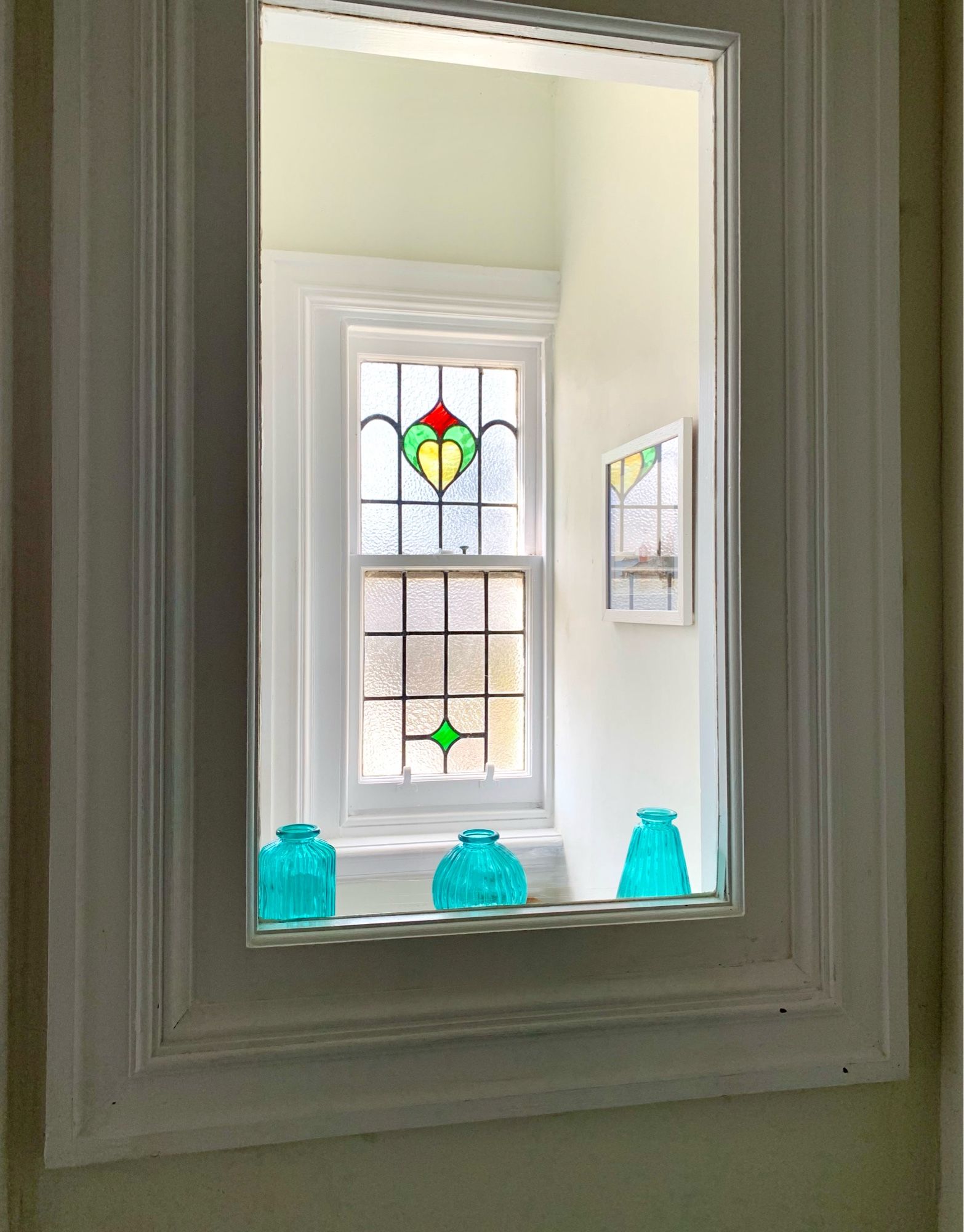 Picture of a stained glass window taken through a clear window, both are framed in white painted wood on cream walls.  The outer window has yellow, green and red stained glass segments with lead mullions and the inner window ledge has 3 turquoise blue decorative bottles.  The overall look is pale creamy white with some primary colour pops.