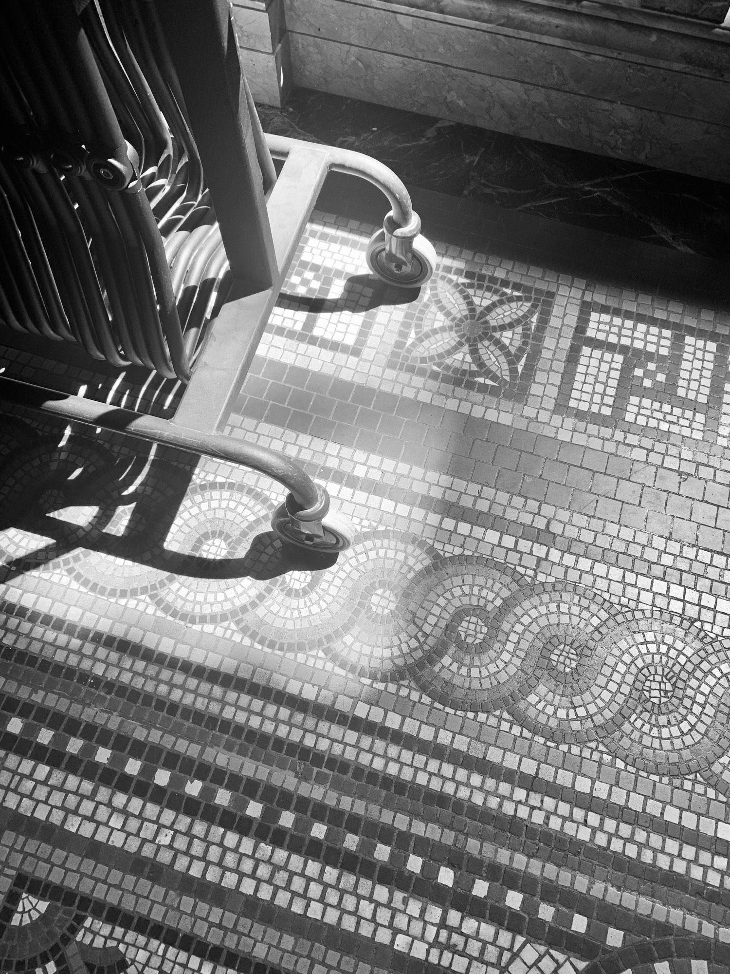 Picture of a beautiful tiled mosaic floor, there is a strong slab of sunshine hitting the trolley wheels of the bank of resting stools